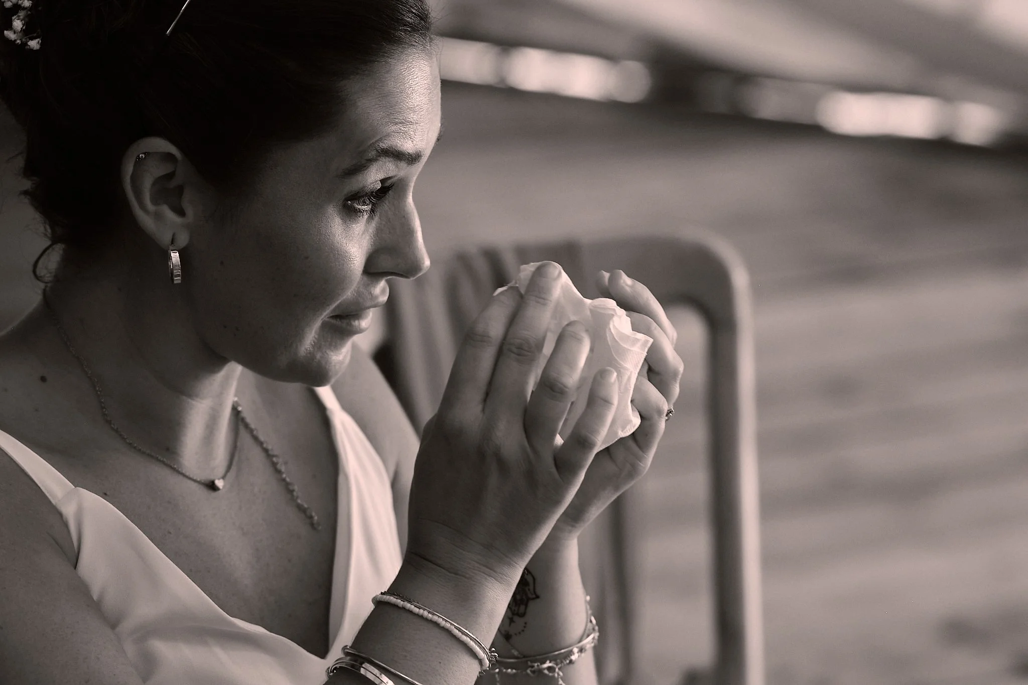 A woman with dark hair tied back, wearing earrings, a necklace, and a sleeveless top, is sitting with her hands clasped near her face, holding a tissue or napkin, in a contemplative pose.