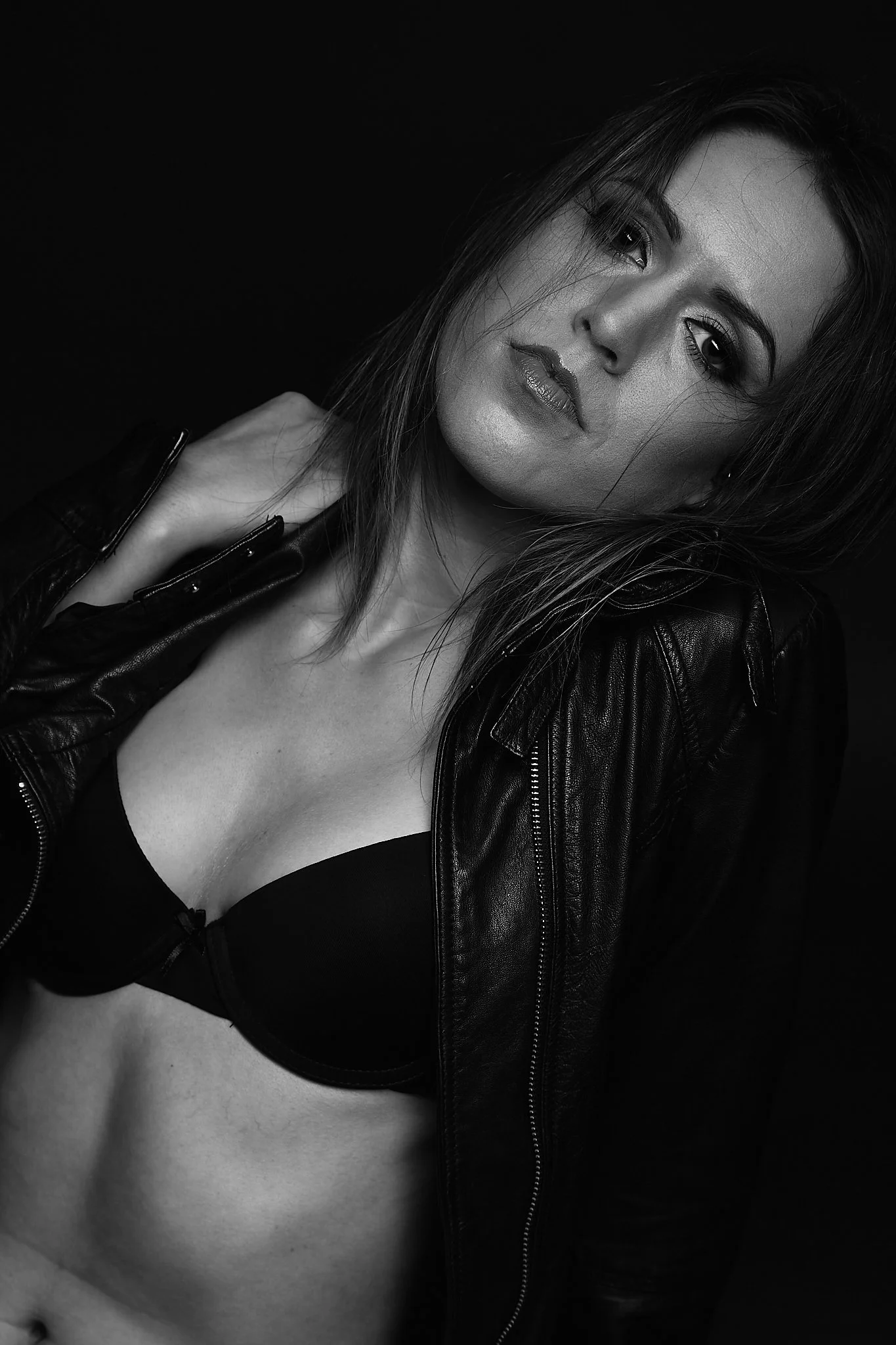 A black and white portrait of a woman wearing a leather jacket and a bra, with her hand on her neck, looking at the camera.