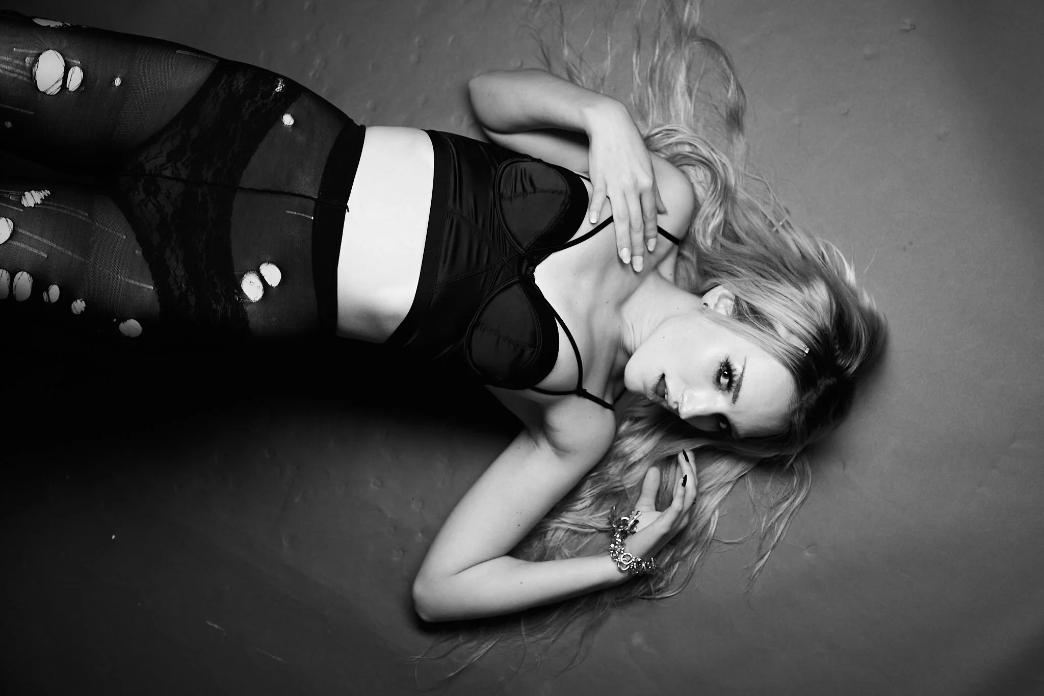 A woman lying on the floor wearing a black lingerie top and ripped fishnet tights with holes, holding her hair with one hand and looking at the camera with a confident expression.