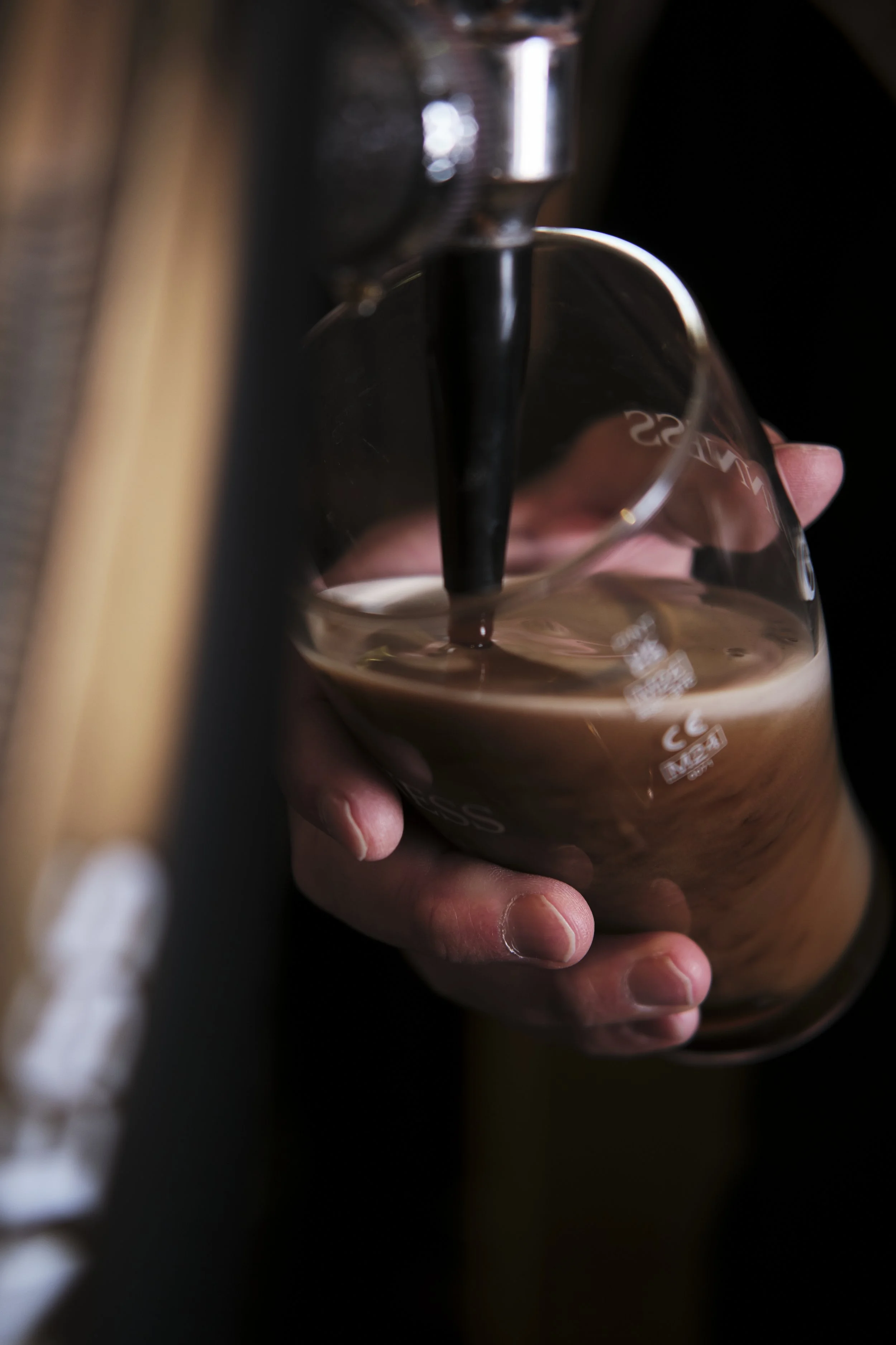 A close-up of a person's hand holding a glass of chocolate milk underneath a chocolate milk dispenser, with the milk being poured into the glass.