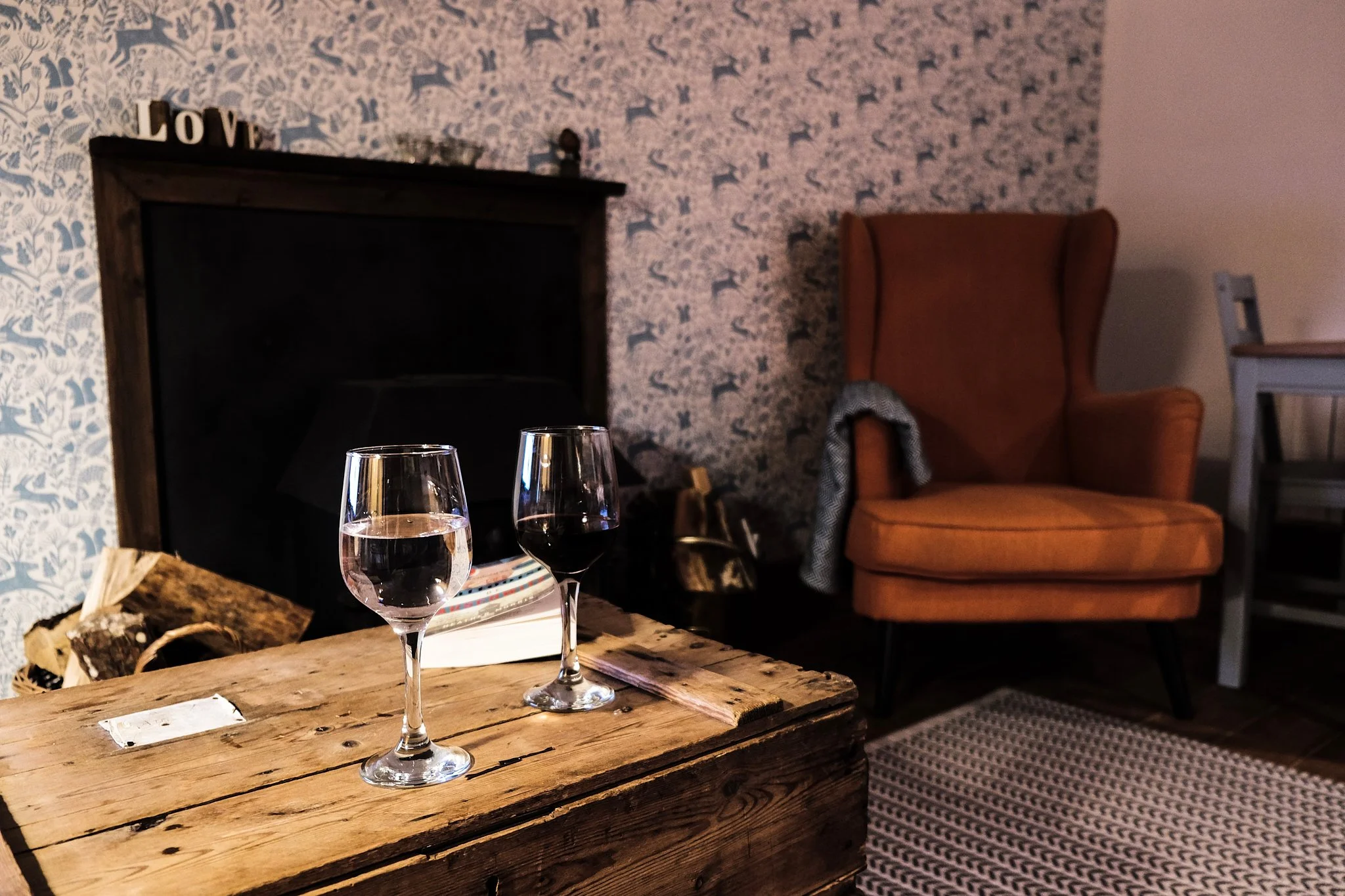 A rustic wooden coffee table with a glass of water, a glass of red wine, and a book, in a cozy living room with patterned wallpaper, an orange armchair with a throw blanket, a fireplace, and a striped rug.