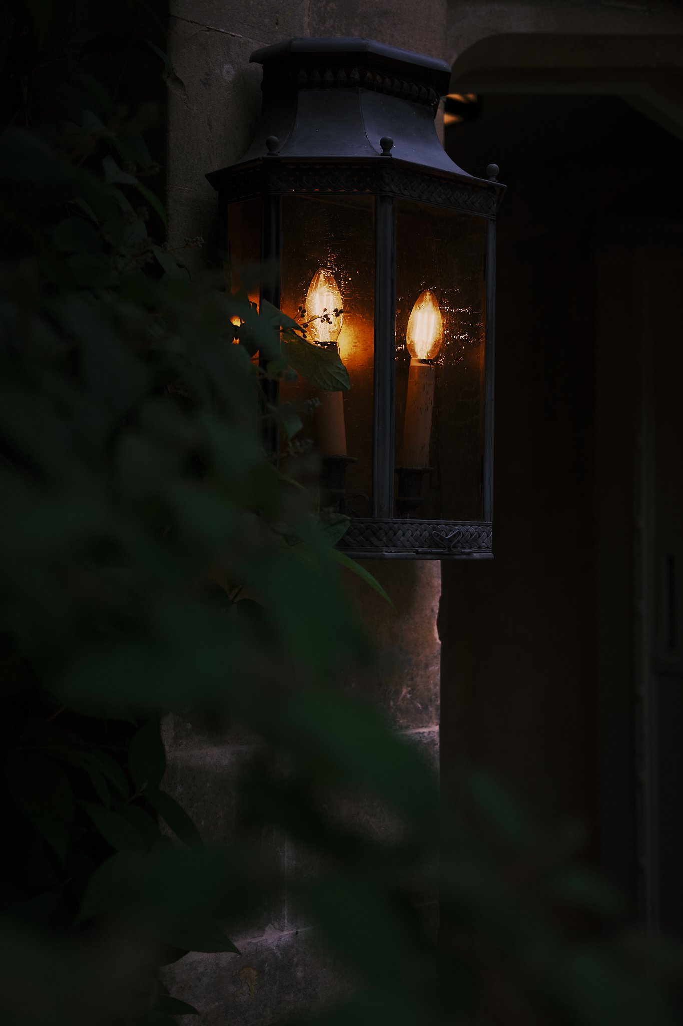 A black outdoor lantern with glass panels, mounted on a wall, with two candles inside illuminating through the glass in warm light, surrounded by dark foliage. Hotel interior photography at THE PIG-in the Cotswolds