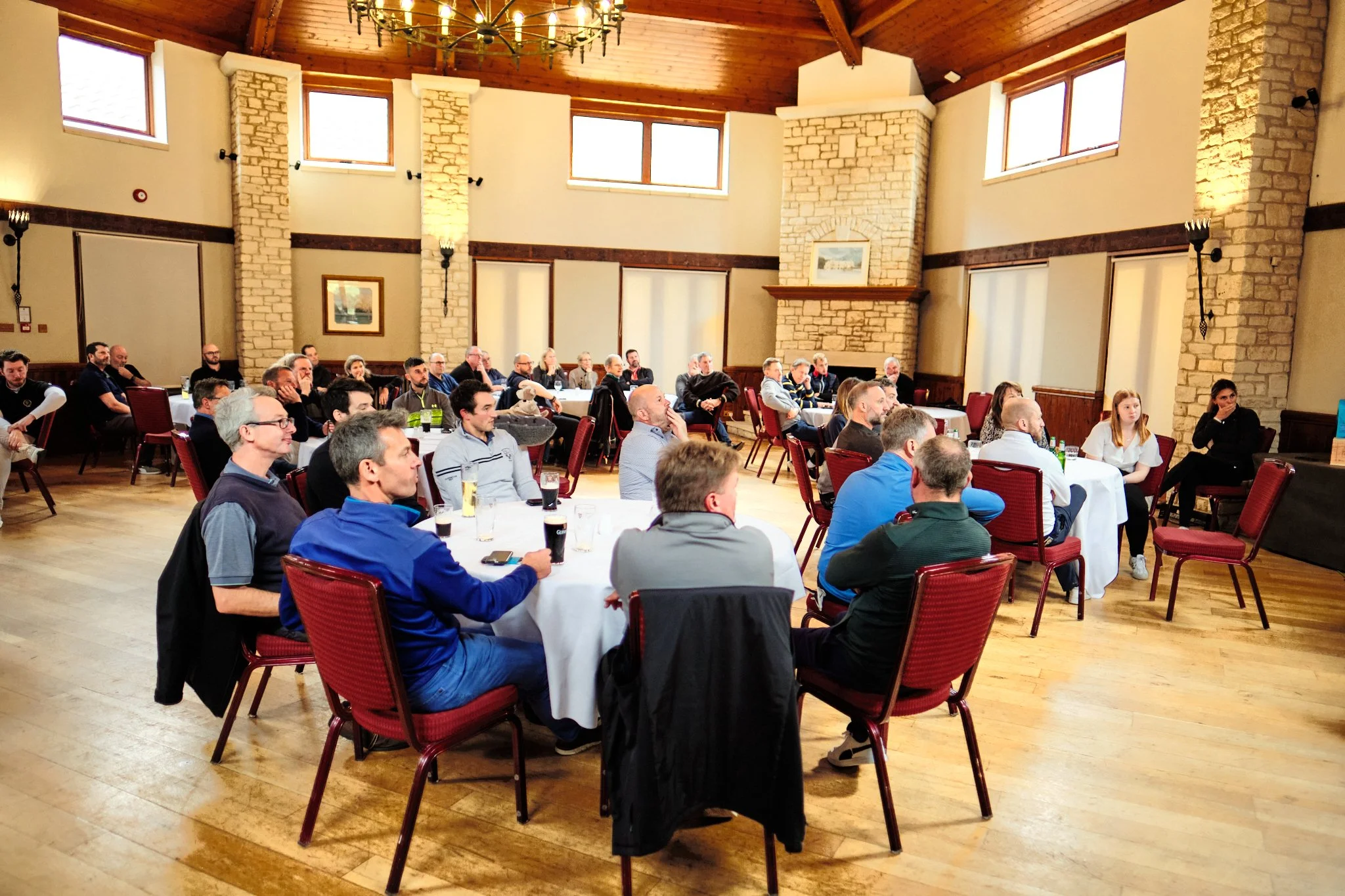 People attending a conference or seminar seated at round tables in a spacious room with wooden floors and stone walls, facing a speaker or presentation area.