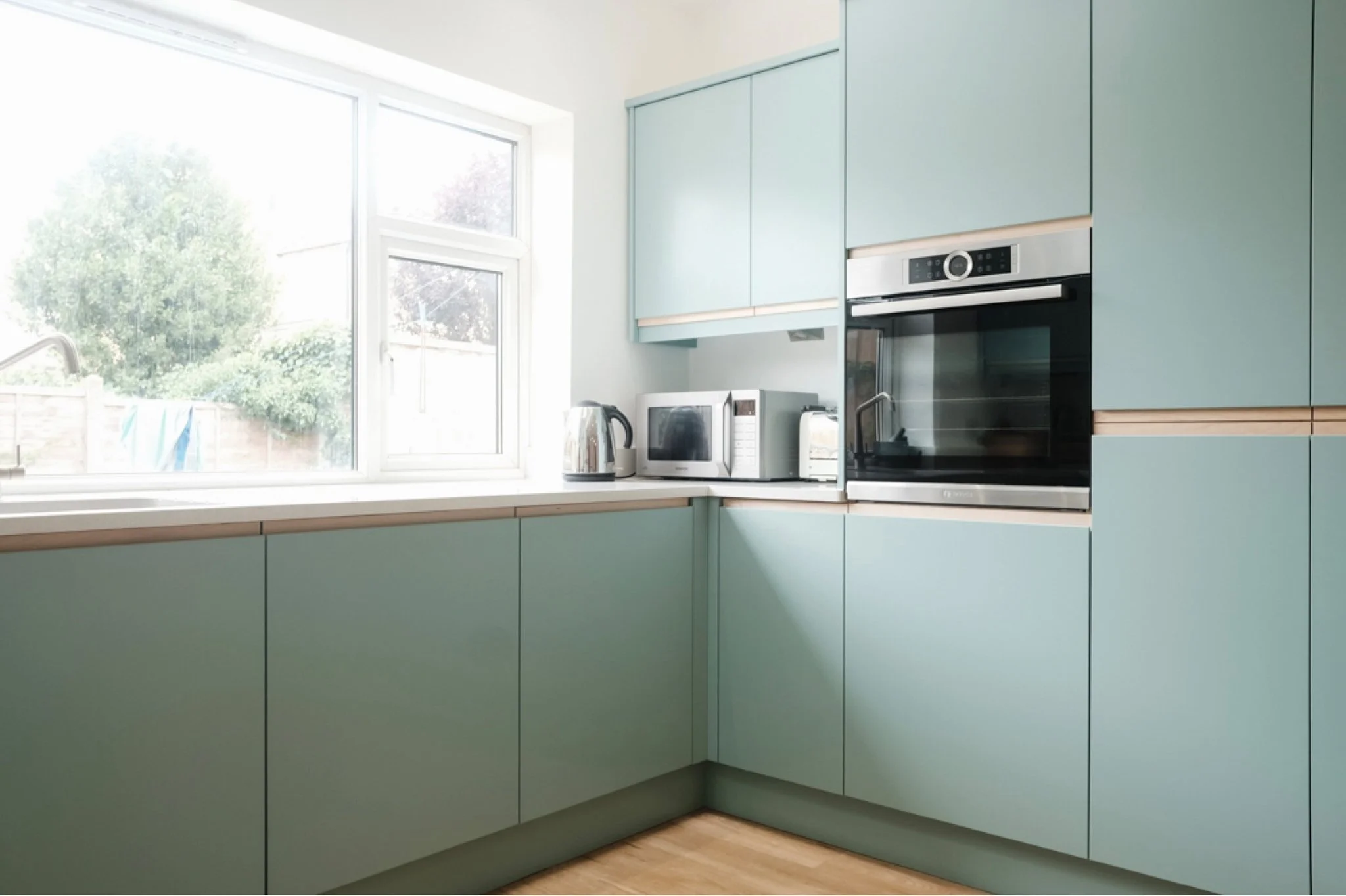 Light blue kitchen cabinets with built-in oven, microwave, and toaster, near a large window with a view of a backyard with trees and a fence.
