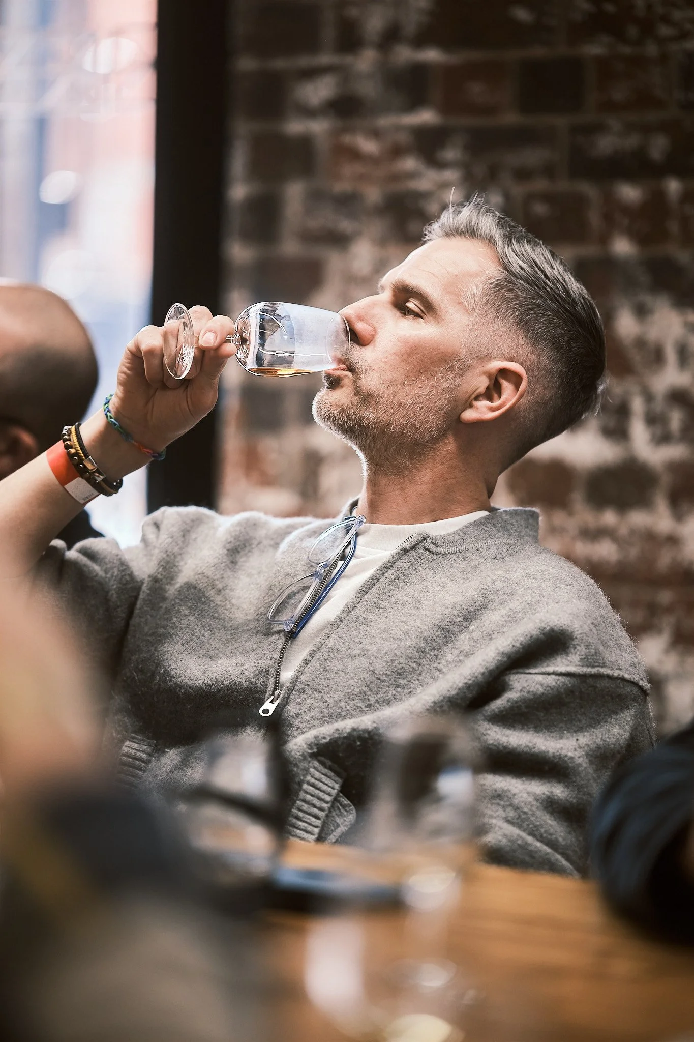 A man in a gray zip-up sweater and white t-shirt drinks from a glass of wine or champagne at a social gathering, with a blurred background of a brick wall and a window.