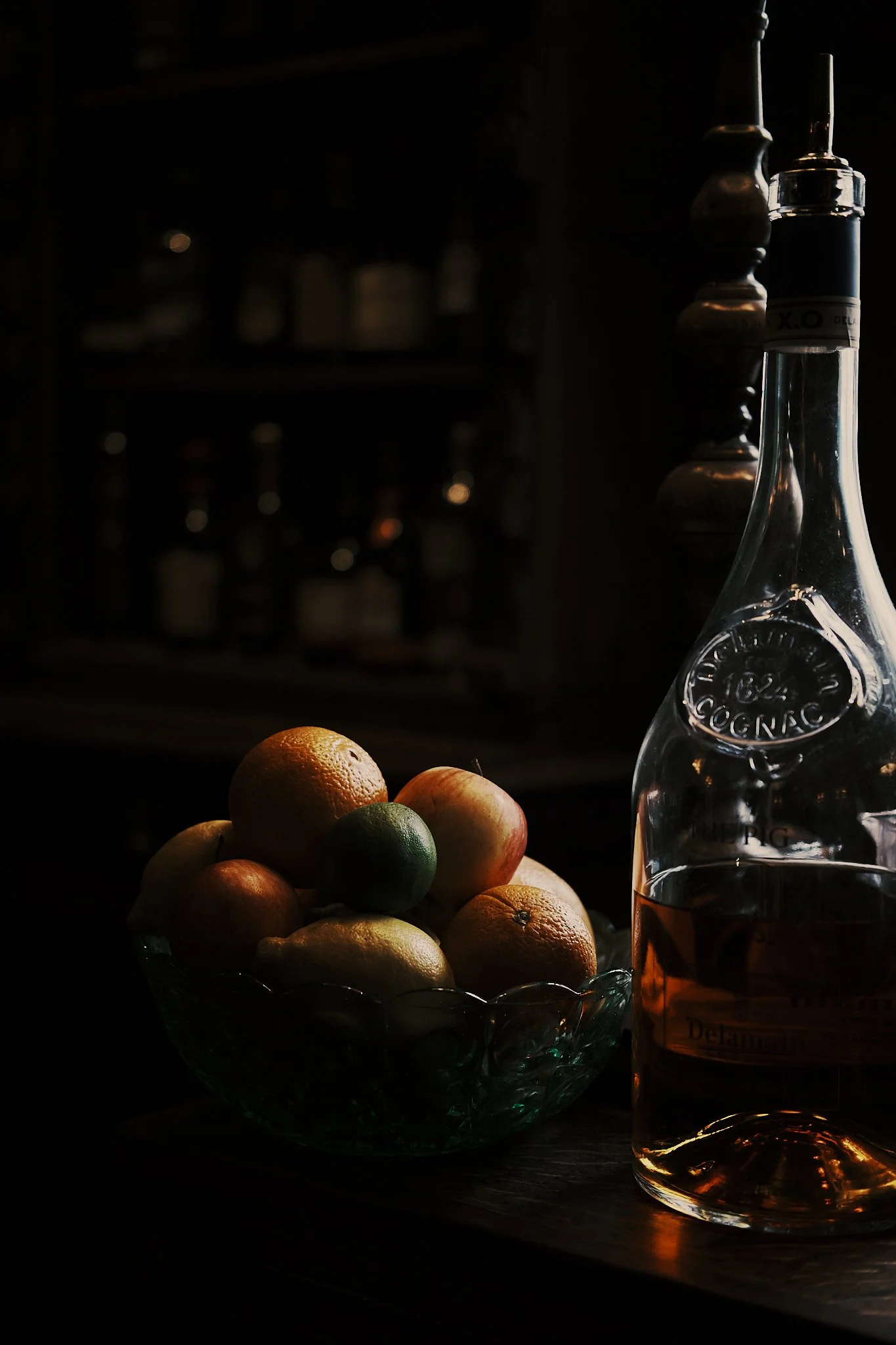 A bowl of citrus fruits including oranges, lemons, and limes with a bottle of liquor beside it, set on a wooden surface in low light. Hotel interior photography at THE PIG-in the Cotswolds