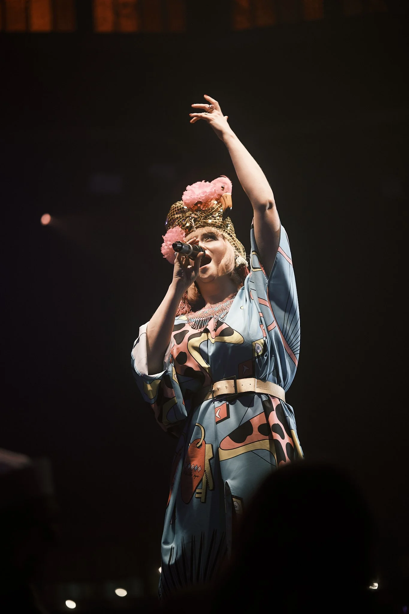 A woman performing on stage, wearing a colorful dress, a hat with pink feathers and gold netting, holding a microphone, raising her hand.