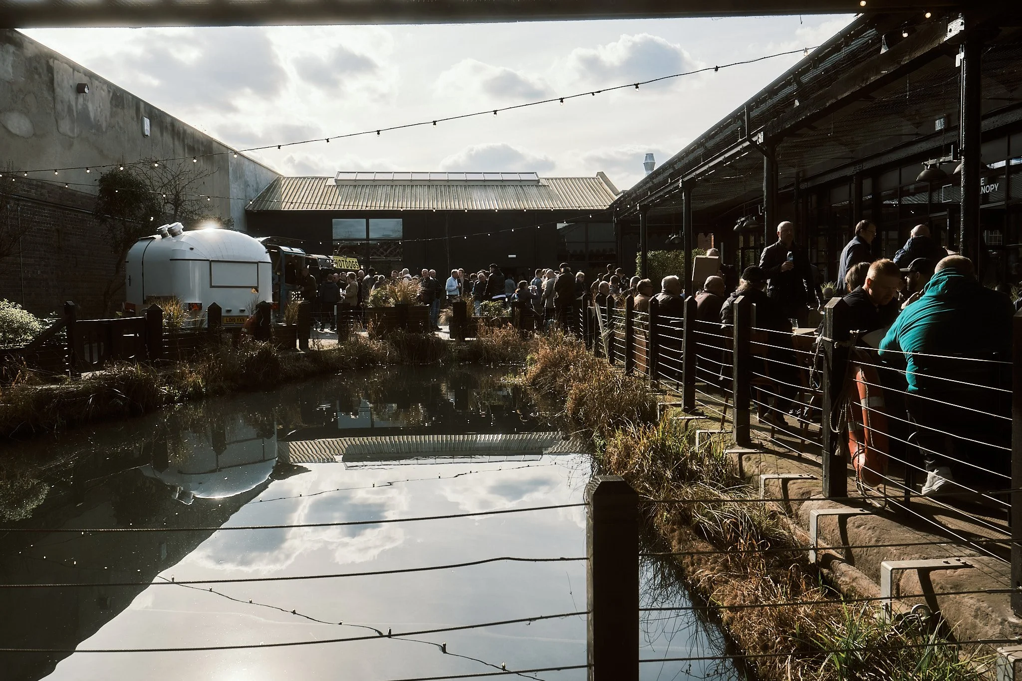 People standing in line along a railing at an outdoor venue, with a pond reflecting the sky and surrounding structures in the foreground.