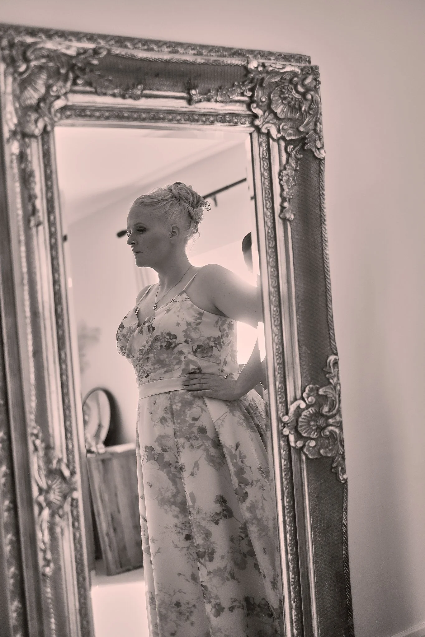 A woman with short, light-colored hair styled in waves, wearing a floral dress, is seen in a mirror with an ornate decorative frame. She has a serious expression and is looking down slightly. The room appears to be a bedroom or dressing area with fur