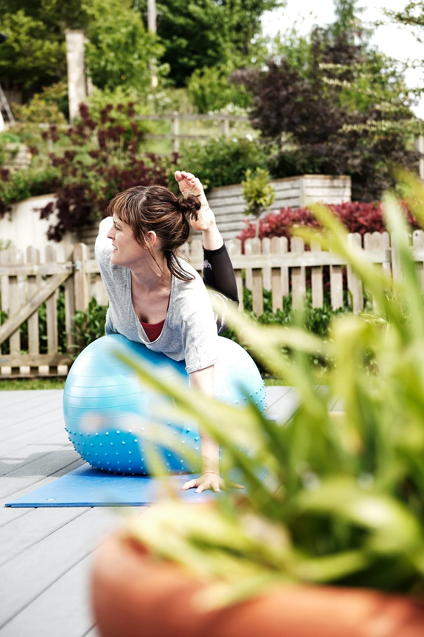 A woman practicing yoga outdoors on a wooden deck, sitting on a yoga mat, using an exercise ball, surrounded by garden plants and trees.