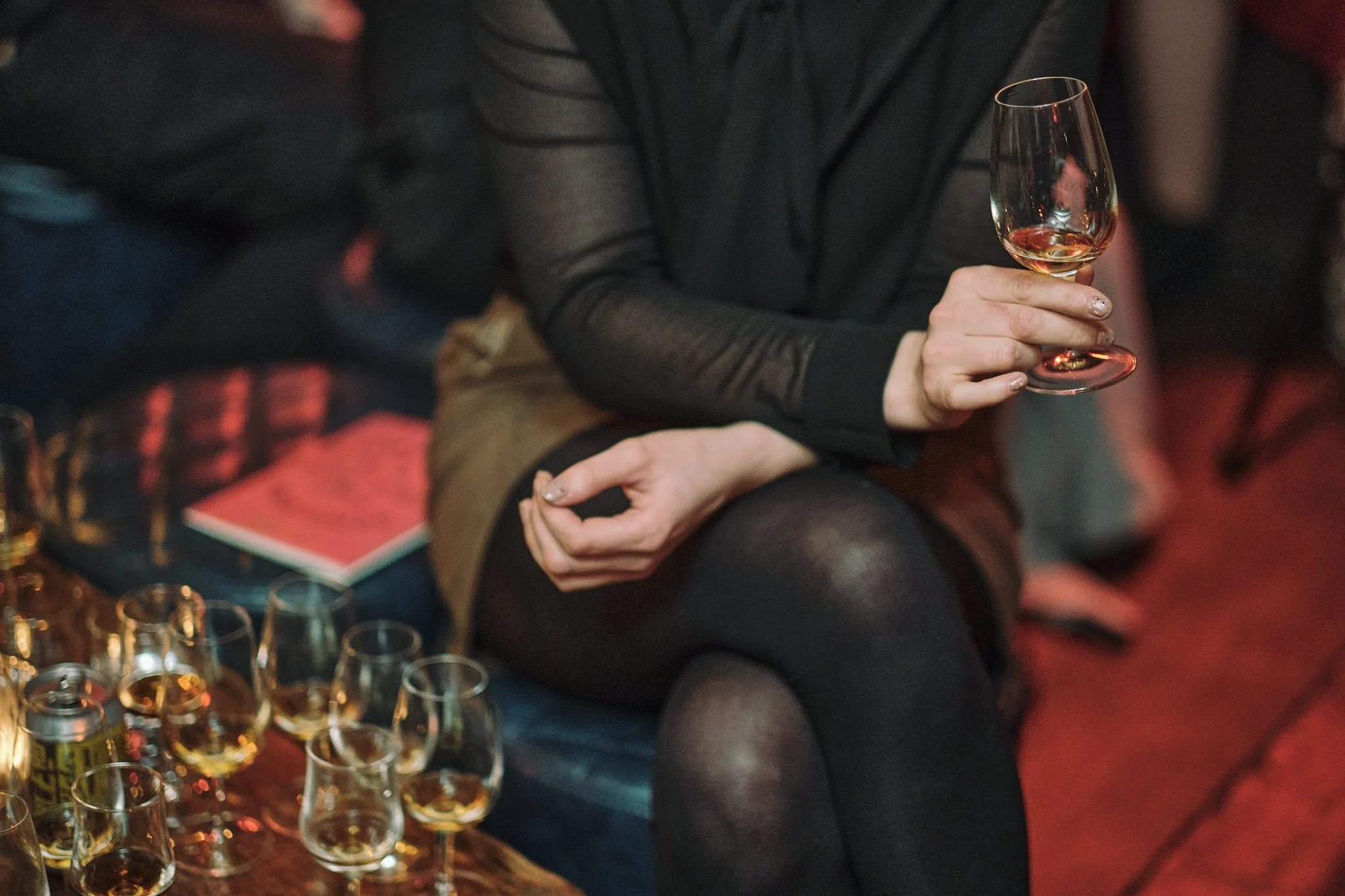 Person sitting on a couch holding a wine glass, with multiple empty and full glasses on the table in front of them.