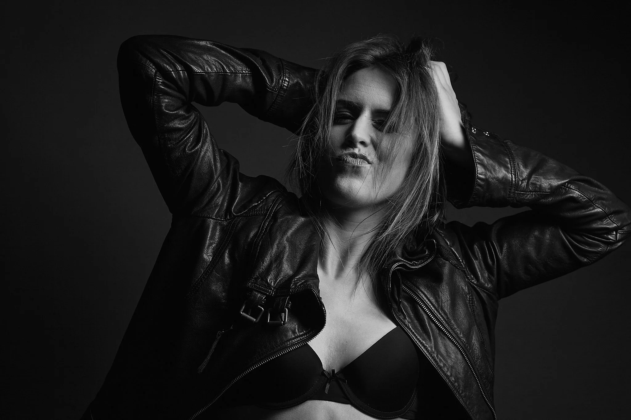 Black and white portrait of a woman with tousled hair, wearing a leather jacket and a bra, with her hands in her hair, posing against a dark background.