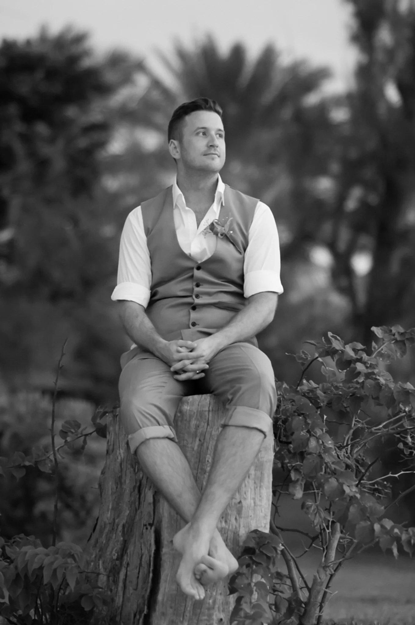 A black and white photo of a young man sitting on a tree trunk outdoors, with trees and foliage in the background.