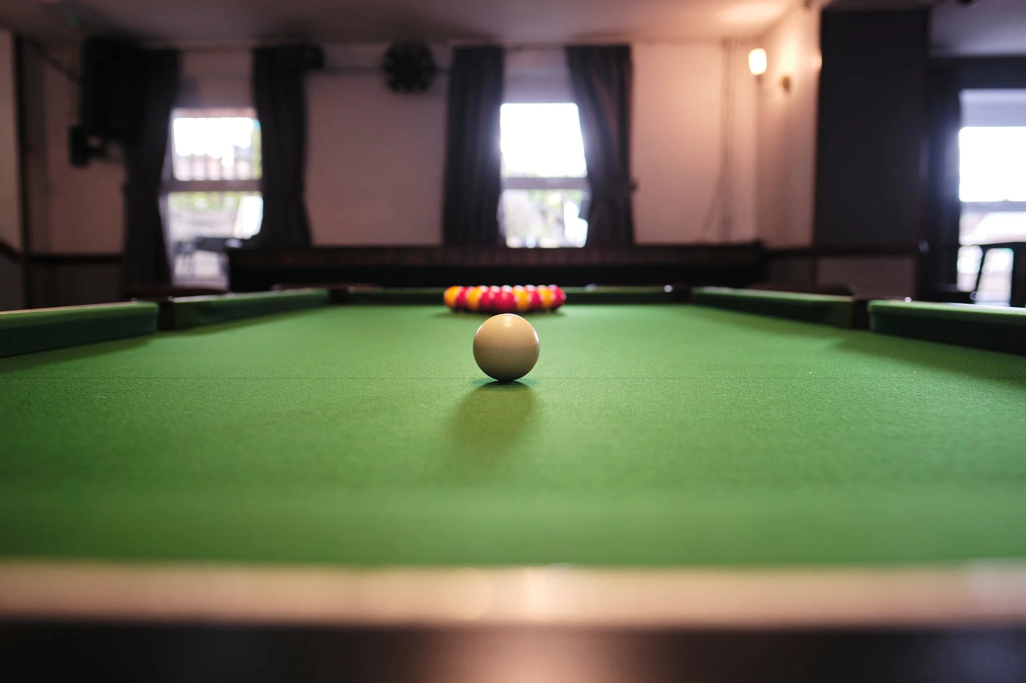 A pool table with a white cue ball centered on the green felt surface, with the rack of colored balls lined up at the far end, in a room with windows and curtains.