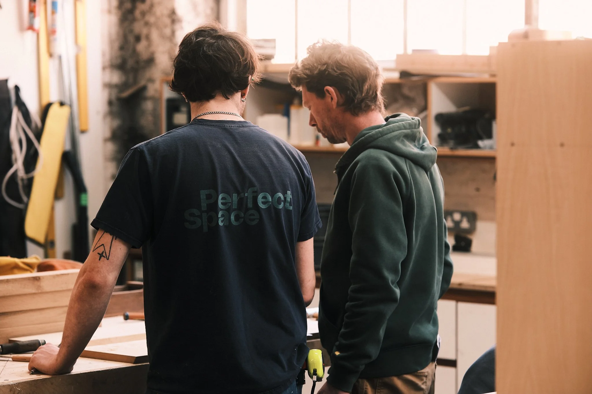 Two people stand close together in a woodworking workshop, examining a project on the workbench.