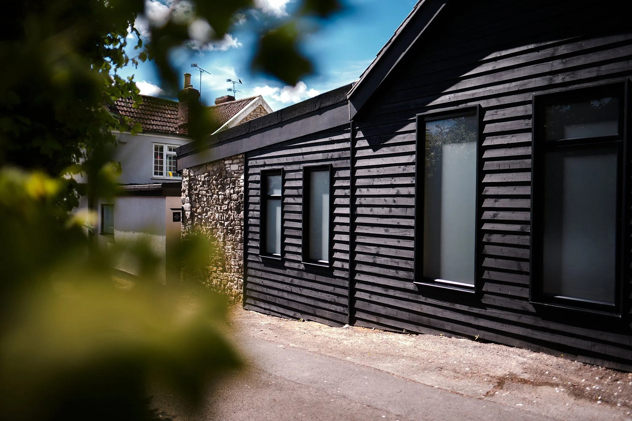 Modern house with black wooden siding and tall rectangular windows, partially obscured by blurred green foliage in the foreground, under a partly cloudy sky.