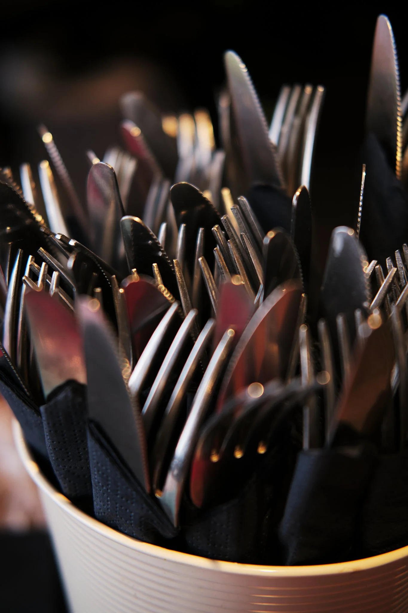 Close-up of a container filled with assorted metal cutlery, including knives, forks, and spoons, arranged upright with a dark background.