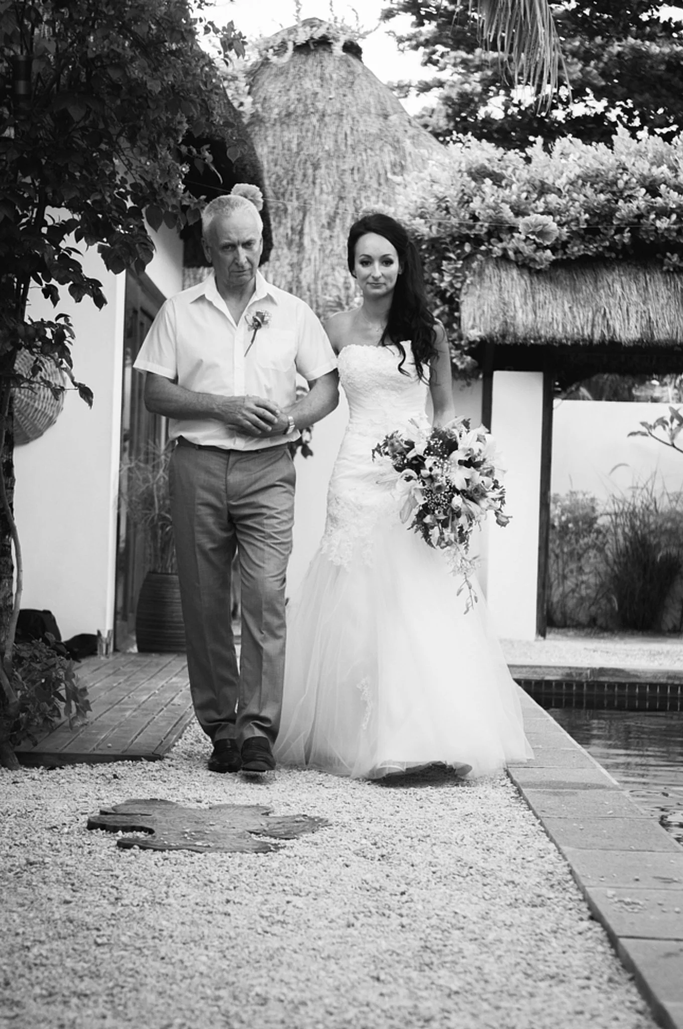 A man and a woman walking together outdoors, the woman in a wedding dress holding a bouquet and the man dressed in a casual shirt and pants, with tropical thatched-roof structures and lush vegetation in the background.