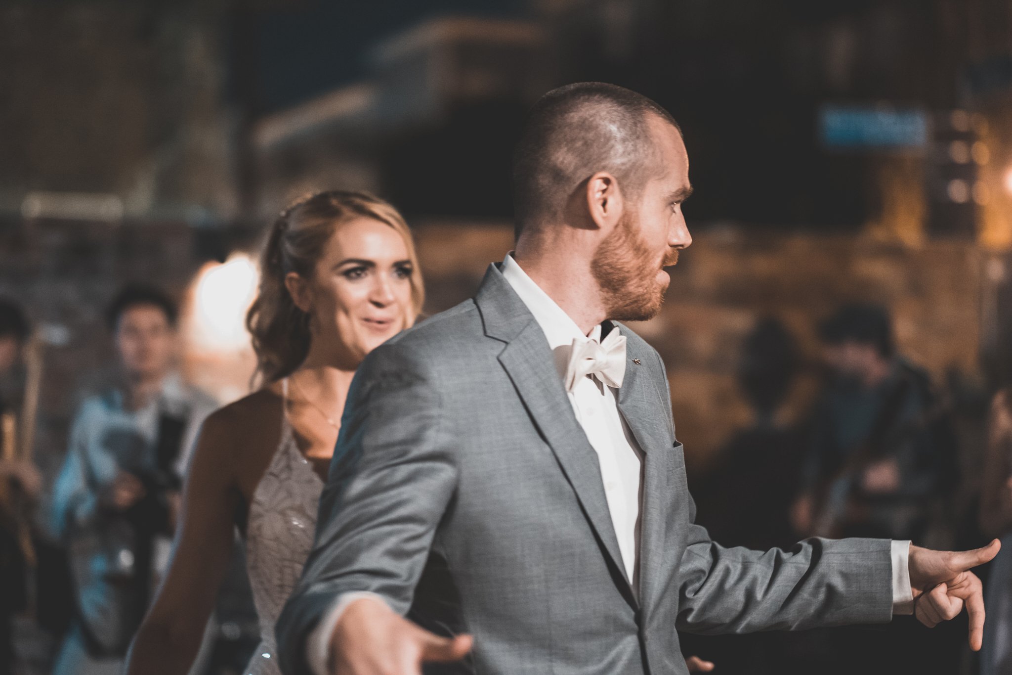 A man in a light grey suit with a white bow tie, standing with his hand raised, appears to be speaking or presenting at an event. Behind him, a woman with light brown hair and a light-colored dress is smiling. The background is dim with other people 