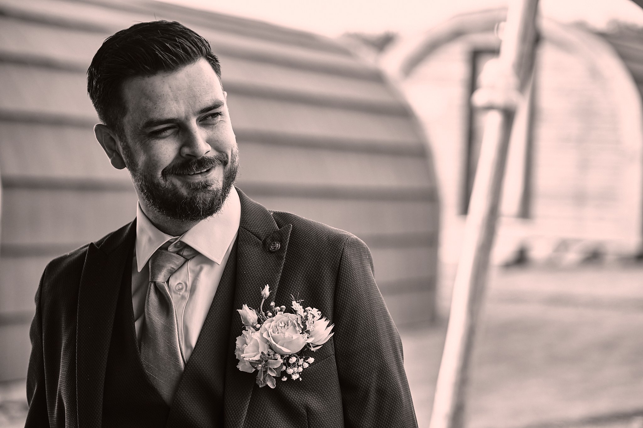 A man in a suit with a boutonniere and a large floral bouquet, smiling and looking to the side, outdoors with a modern architectural background in black and white.