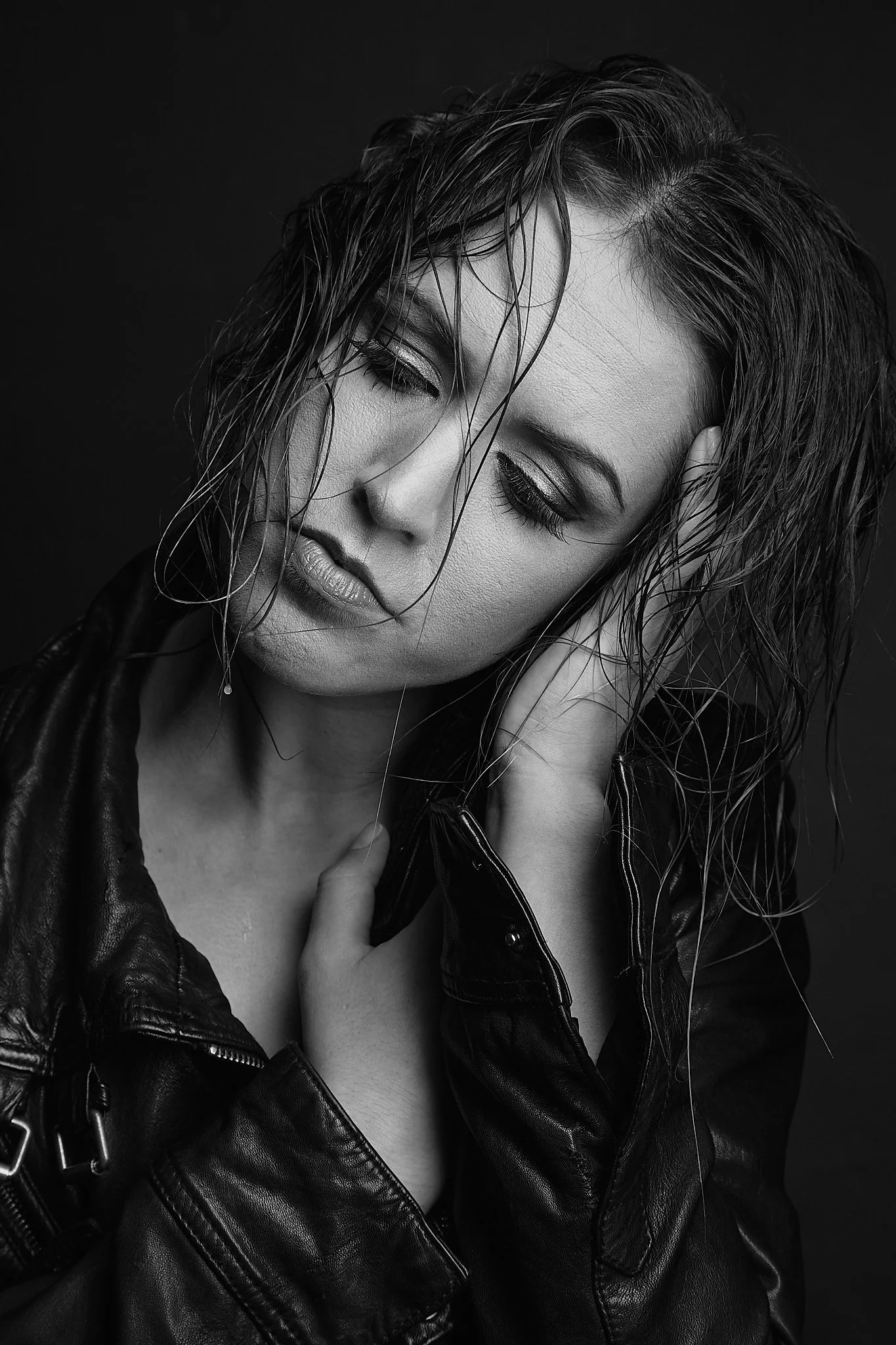 A black and white portrait of a woman with wet hair, wearing a leather jacket, with her eyes closed and hand on her neck.