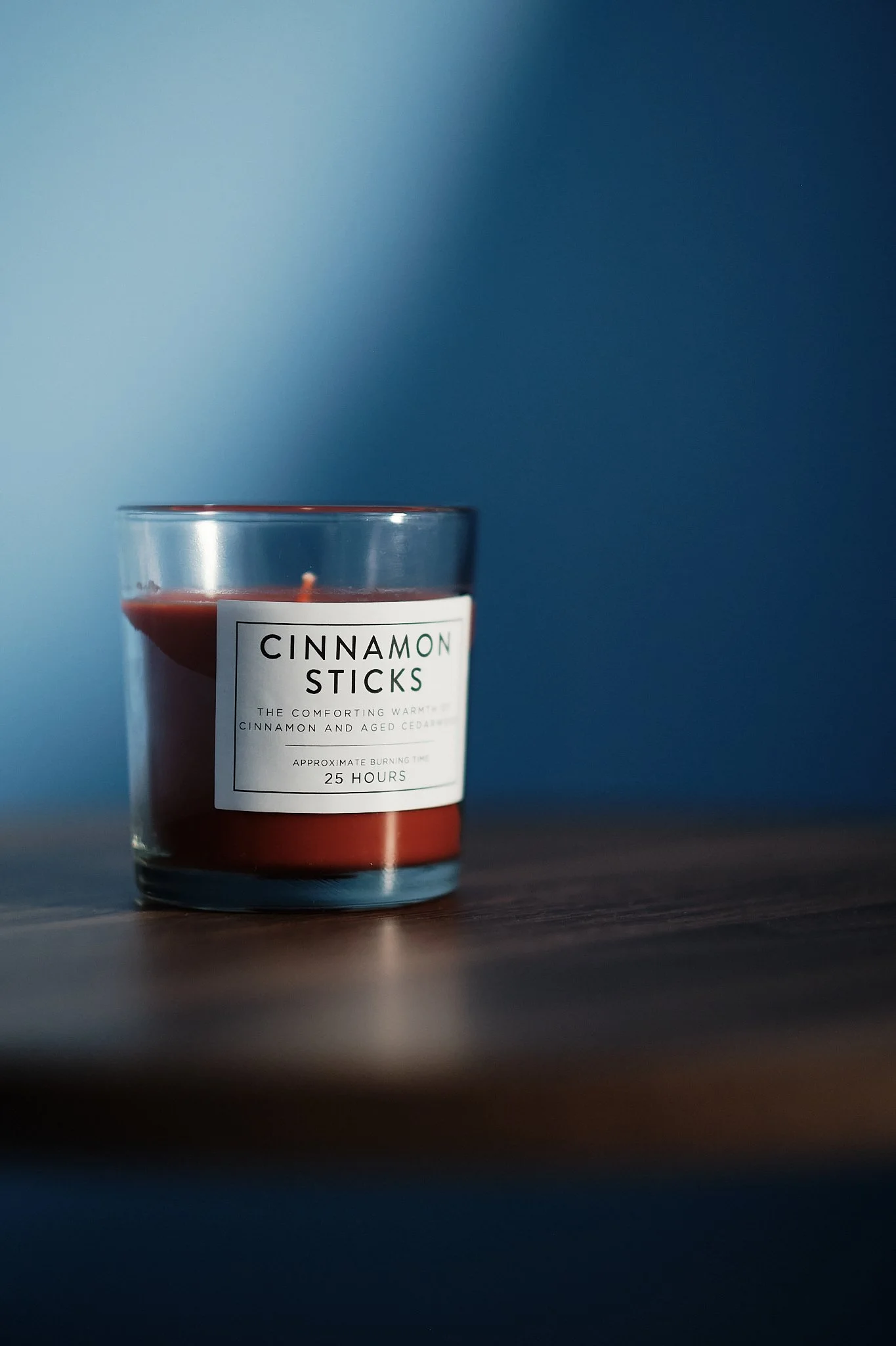 A scented candle in a glass jar with a label that reads 'Cinnamon Sticks' placed on a wooden surface with a blurred gradient blue background.