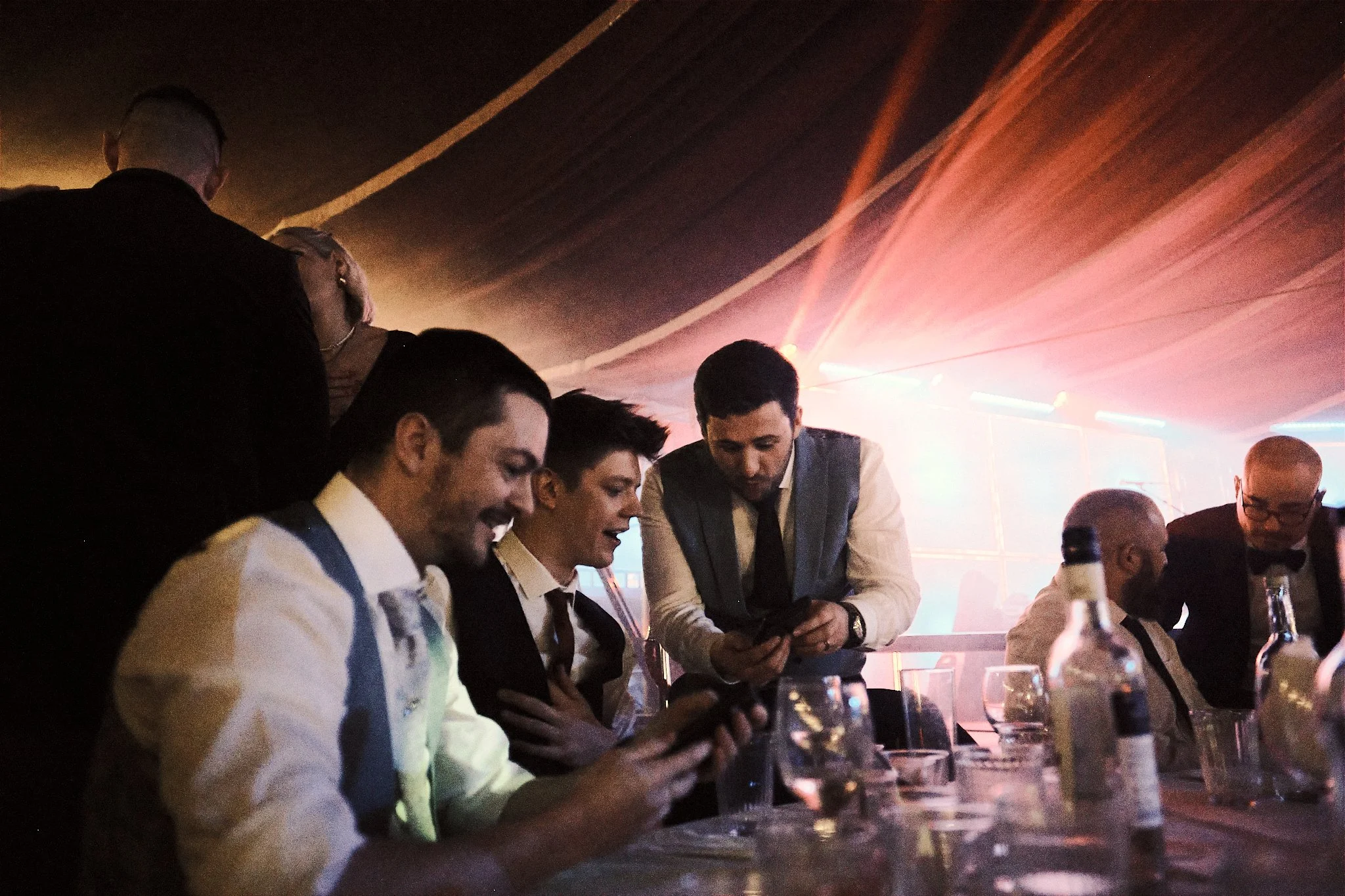 A group of men dressed in formal attire, including tuxedos and vests, gathered around a table at a party or event in a dimly lit room with colorful lights. Some are looking at their phones, and others are engaged in conversation.