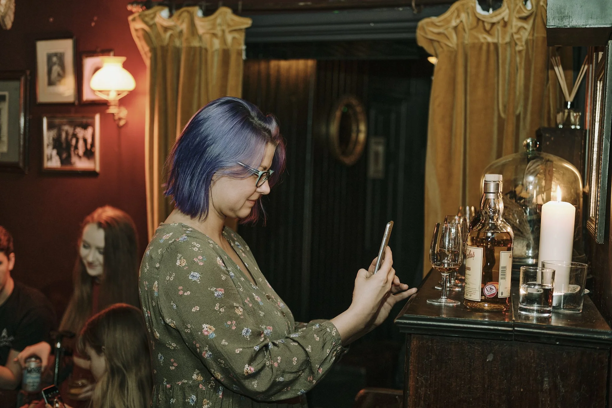 A woman with shoulder-length purple hair and glasses standing at a bar counter, looking at her phone. The bar features a bottle of liquor, a glass of wine, a chalkboard menu, and a lit candle. There are other patrons sitting in the background, and th