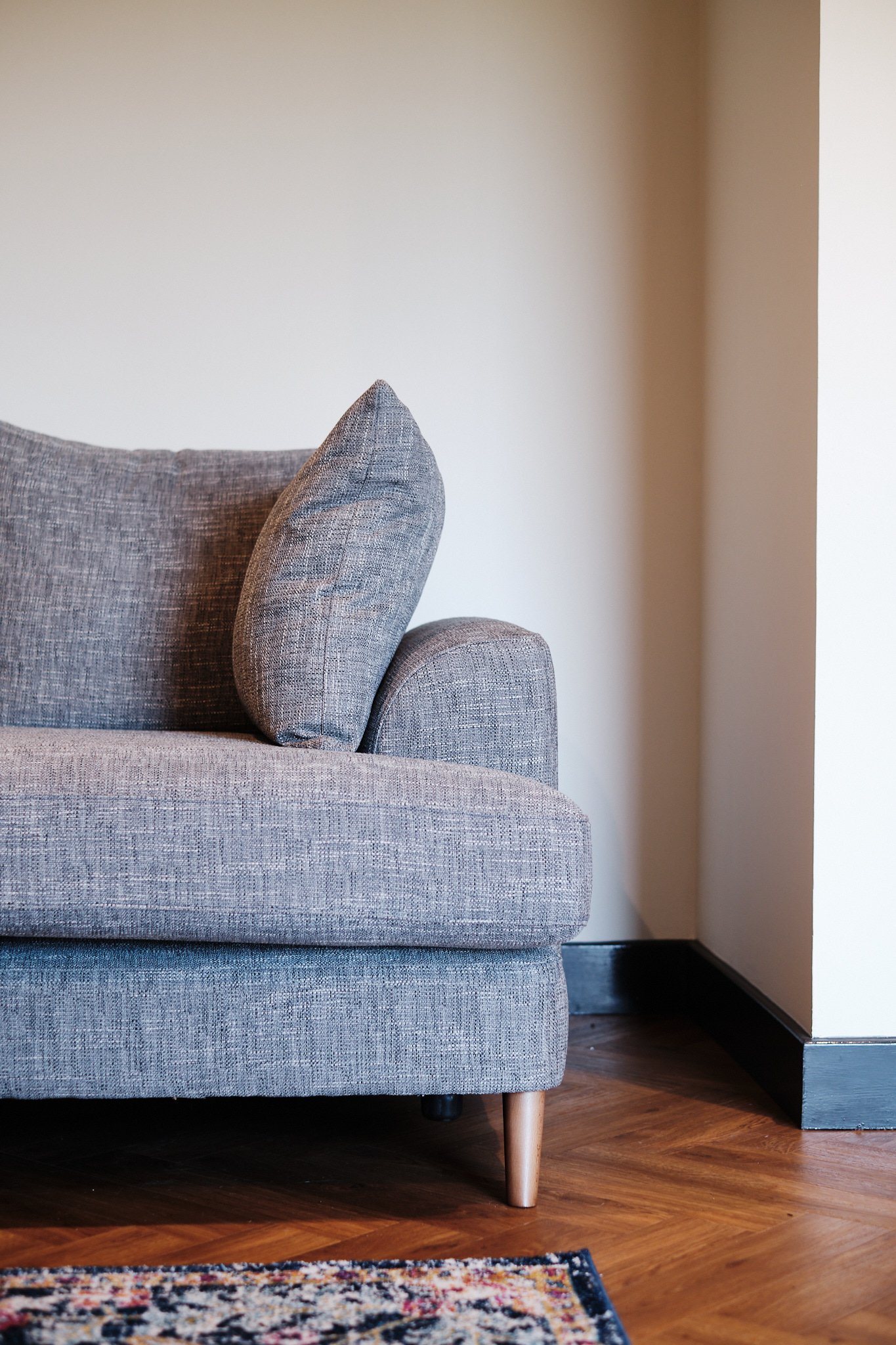 Close-up of a grey fabric sofa with a cushion, wooden legs, in a room with hardwood floor and beige wall.