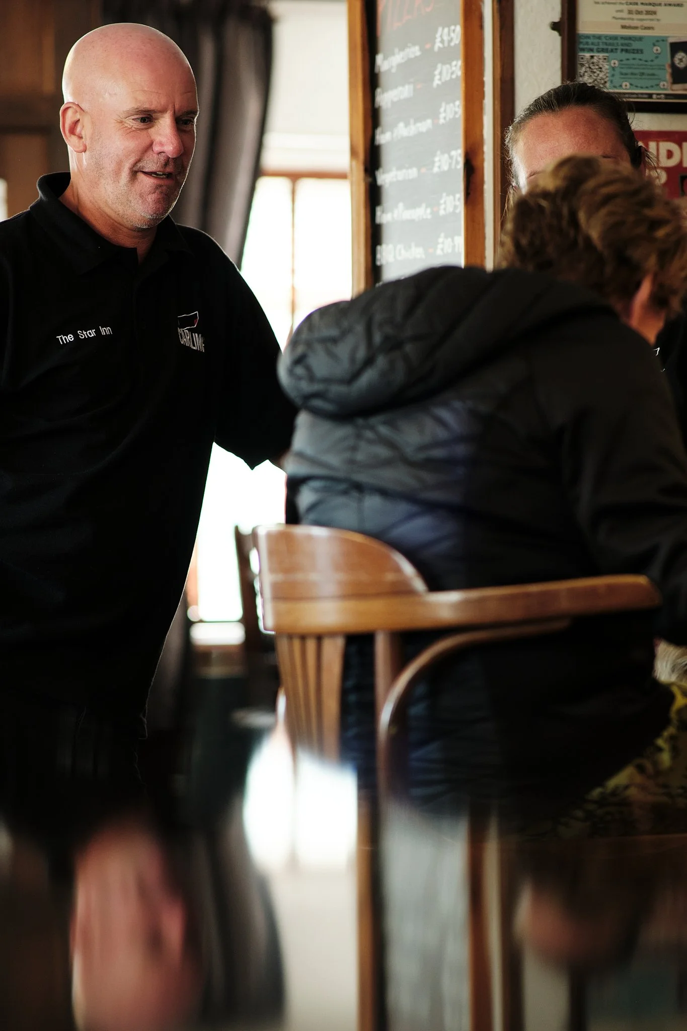 A man with a shaved head and a beard wearing a black polo shirt standing and talking to a seated woman in a black jacket inside a restaurant with a chalkboard menu in the background.