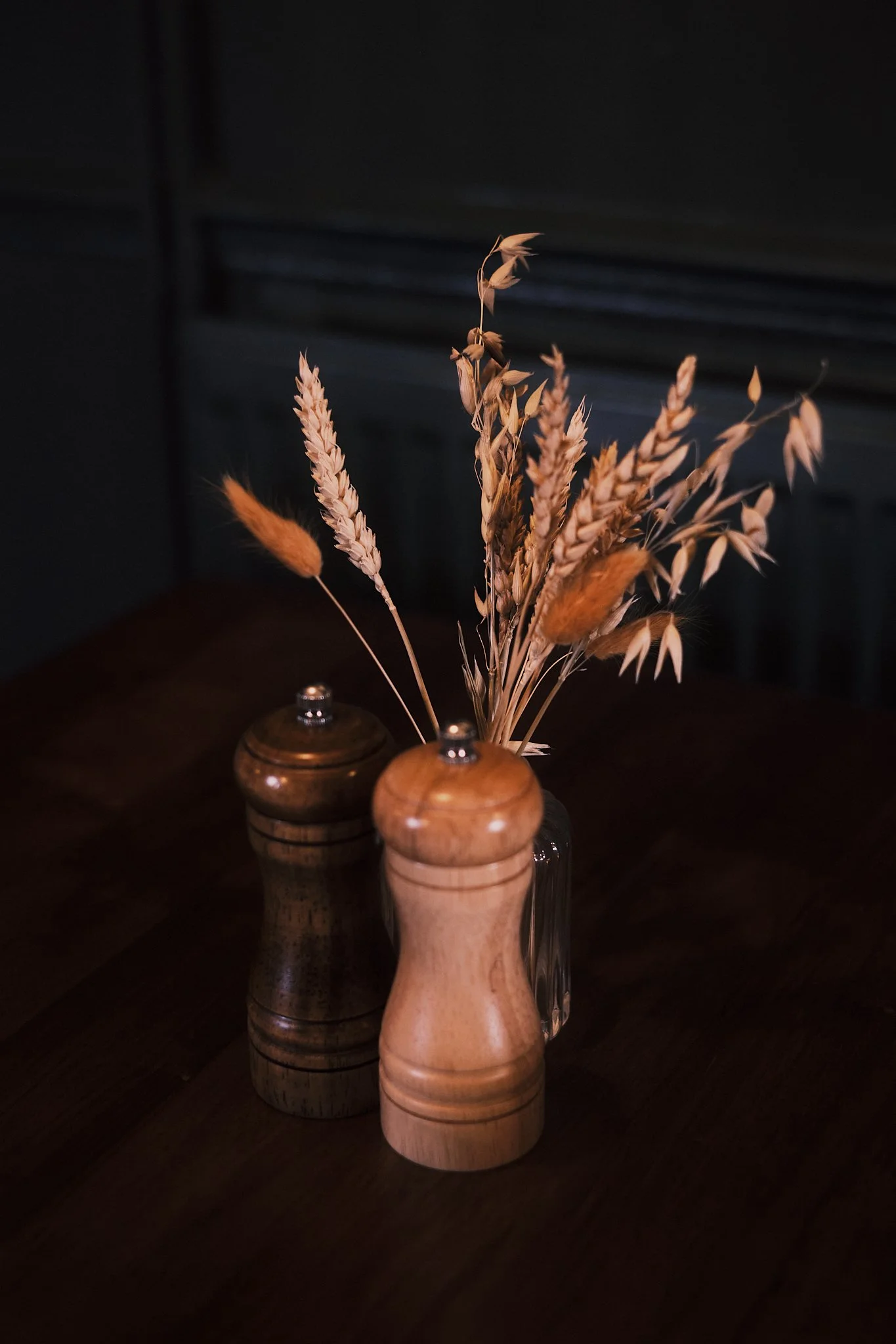Wooden salt and pepper shakers on a dark wooden table with dried flower bouquet in the background.