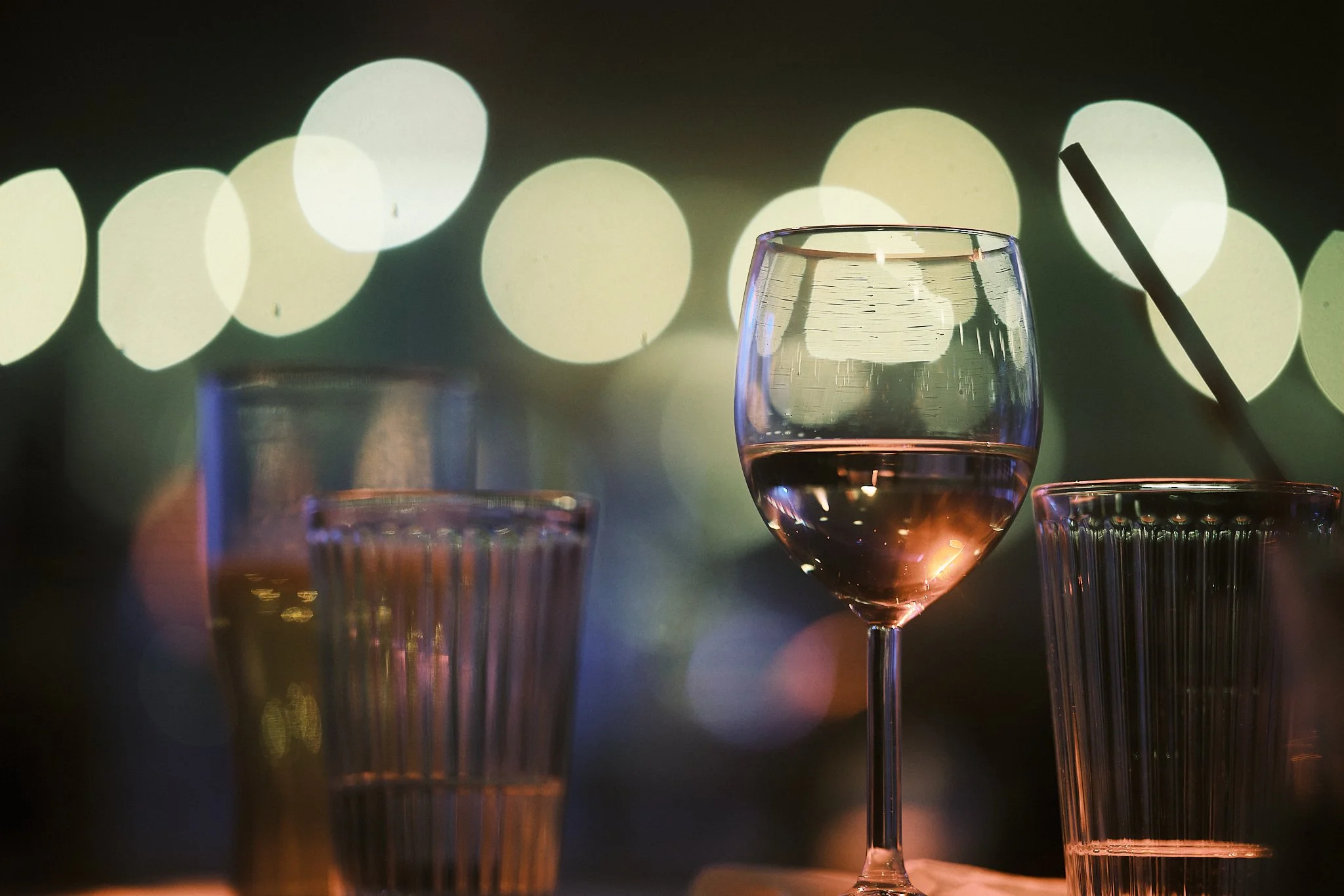 A glass of pink wine, two empty glasses, and a straw in a drinking glass on a table with blurred colorful lights in the background.