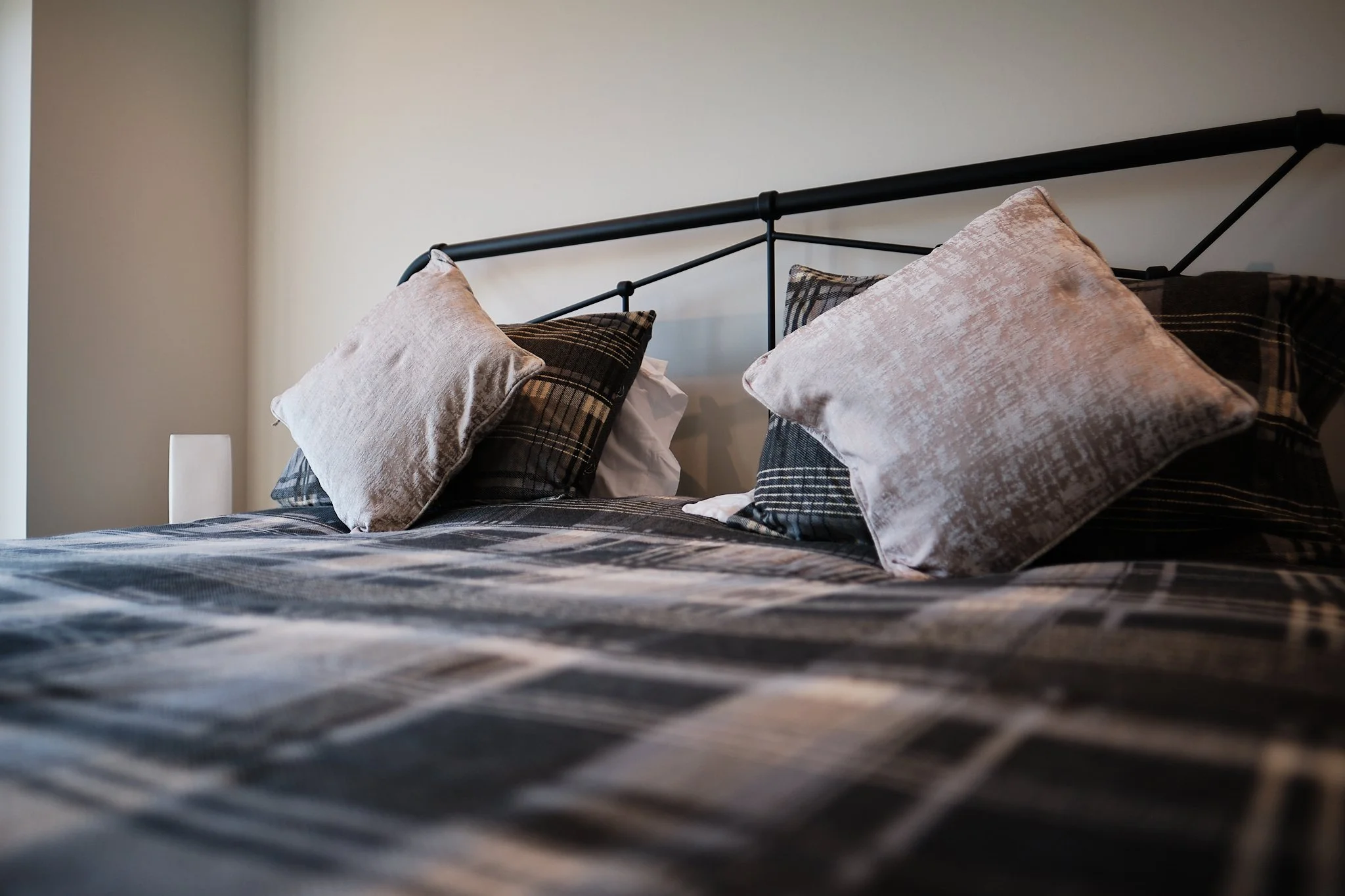Close-up of a neatly made bed with black metal headboard, plaid bedspread, and assorted pillows in beige and black patterns.