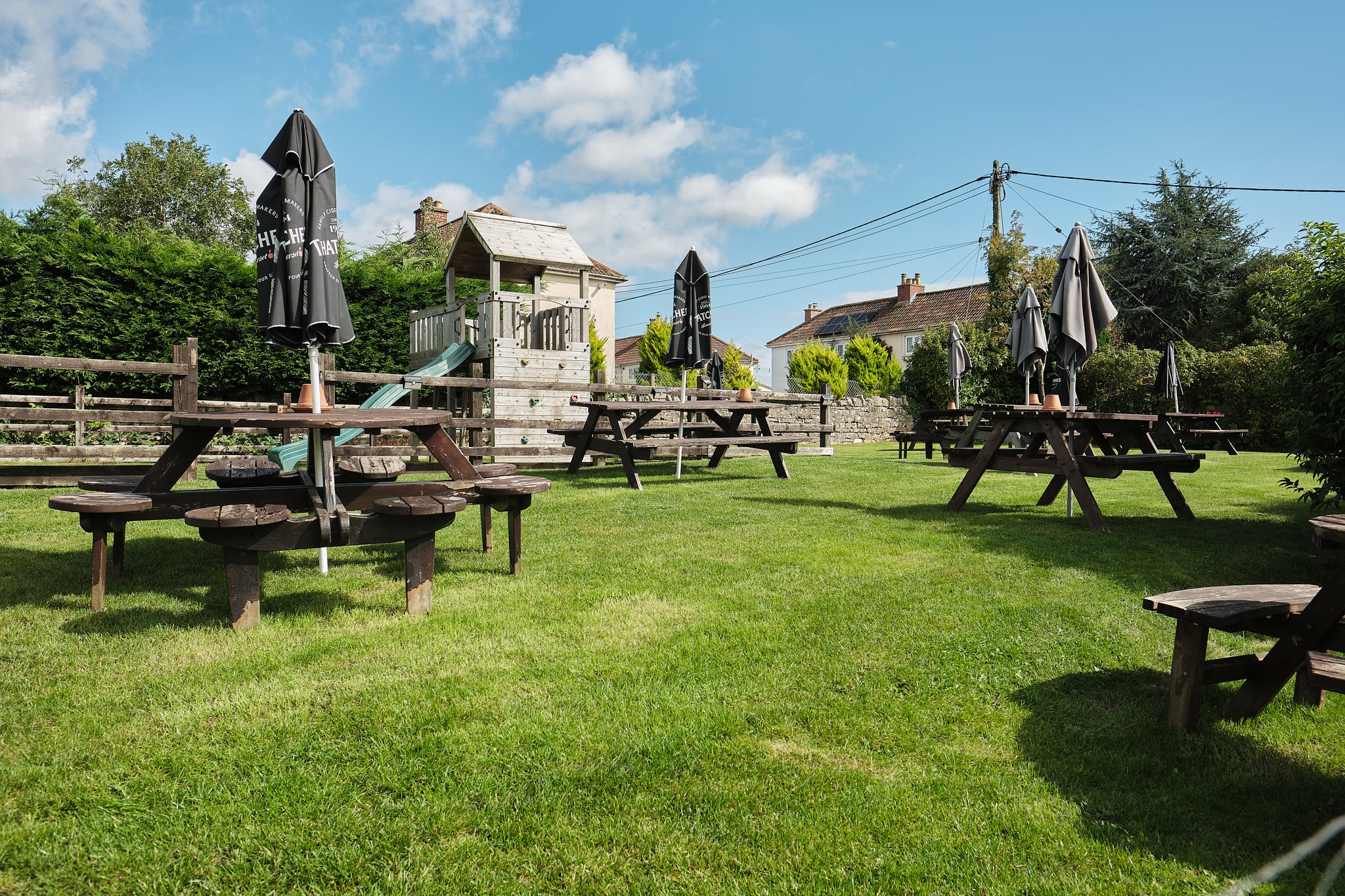 Outdoor garden area with wooden picnic tables and closed umbrellas, green grass, small playhouse, and houses in the background under a partly cloudy sky.
