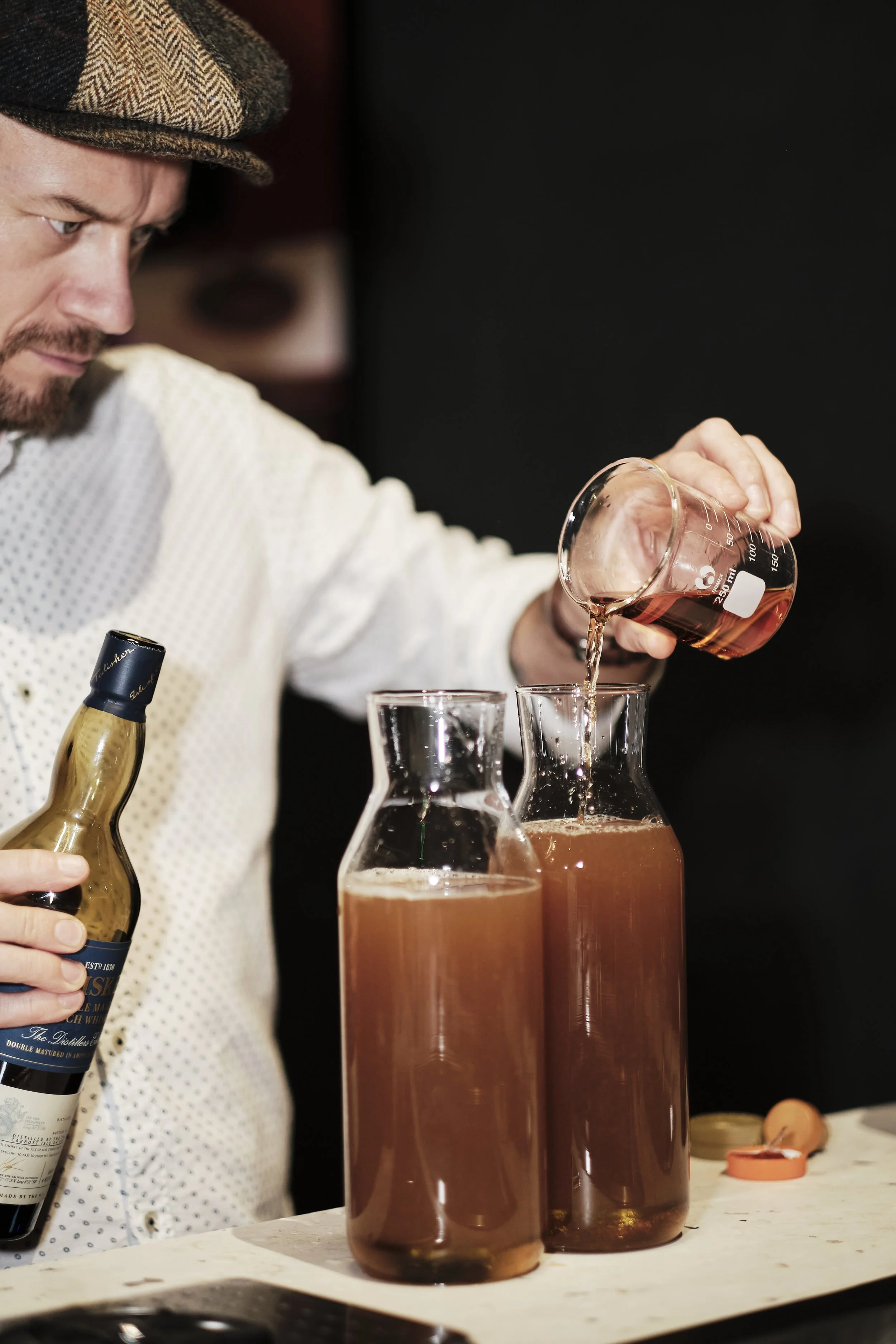 A man in a brown hat and white shirt pouring a brownish liquid into one of two tall glass pitchers, with a bottle in his hand. The pitchers are on a white surface with a black background.