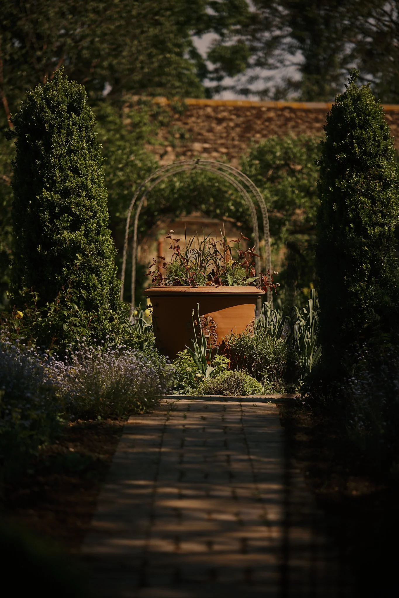 A garden pathway leading to a large flowerpot with plants, flanked by tall, trimmed bushes, with trees and a roof in the background. Hotel interior photography at THE PIG-in the Cotswolds