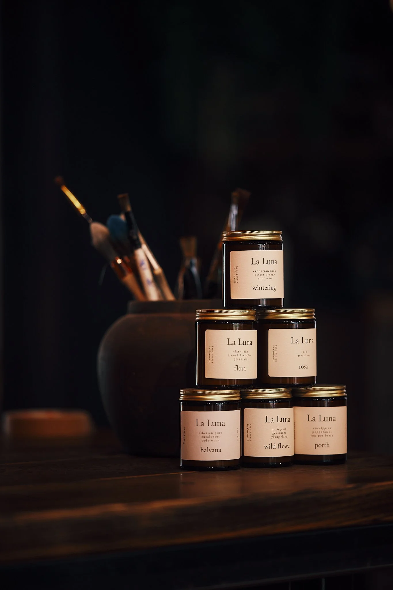 A stack of six jars of La Luna scented candles on a wooden surface, with a dark background and a vase with paintbrushes behind them.
