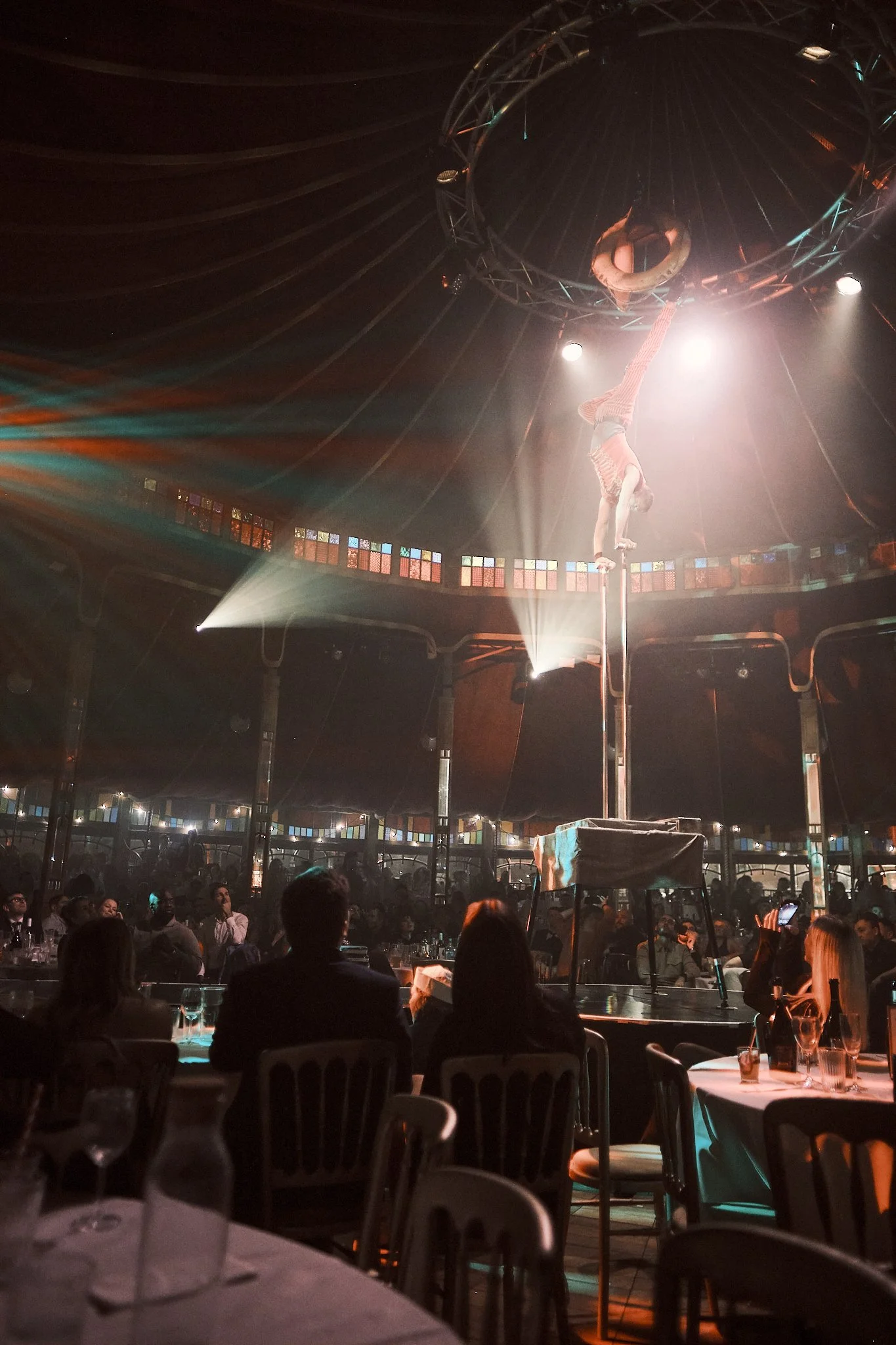 Cirque du Soleil performer on a pole performing an acrobatic trick inside a circular stage, with audience seated around the stage, illuminated by stage lights.