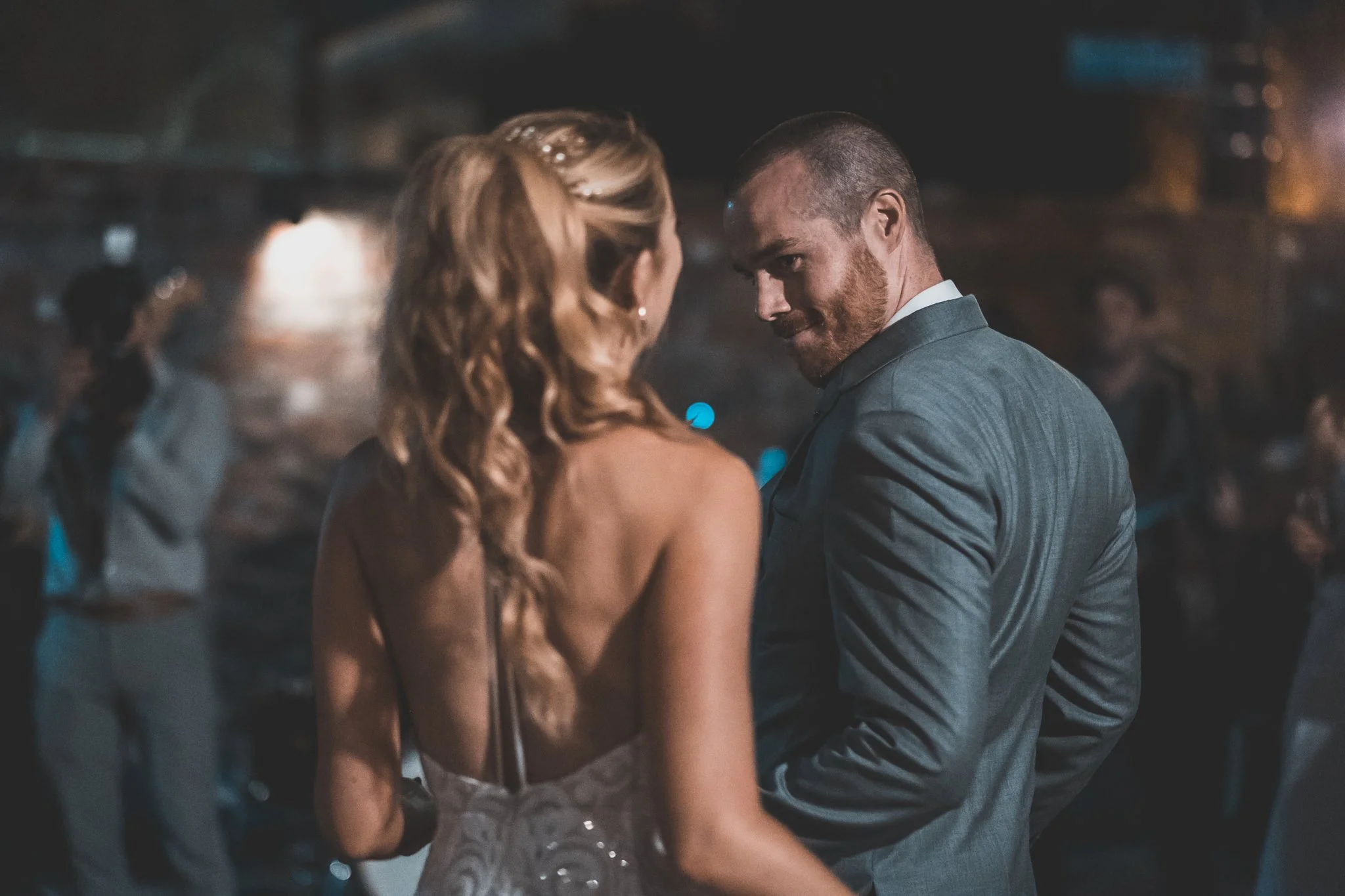A couple dancing closely at a social event, with a woman with curly red hair in a dark dress and a man with a beard and short hair in a suit, in a dimly lit environment.