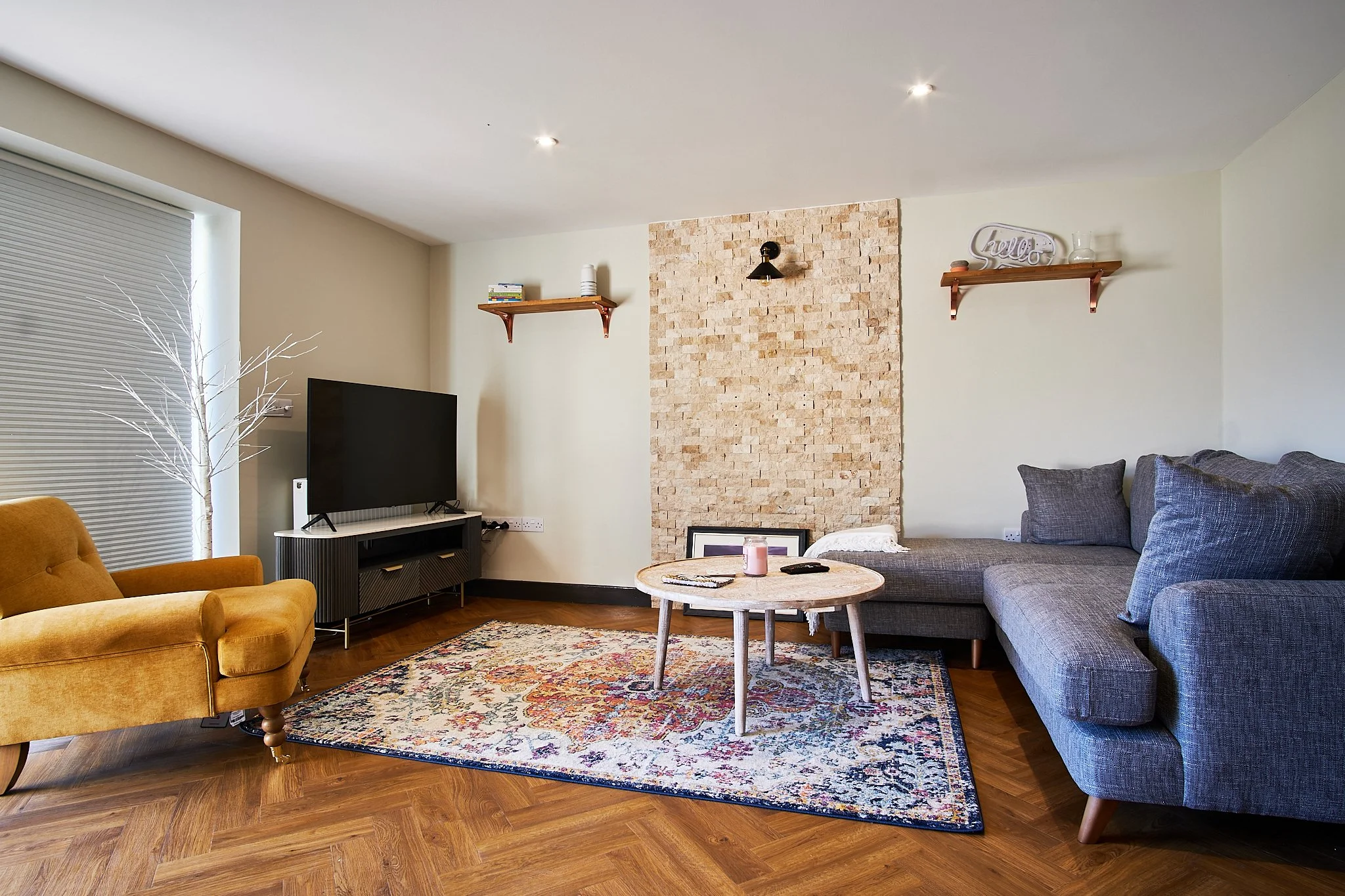 Living room with yellow armchair, gray sectional sofa, TV on black stand, and wooden shelves on wall, on a patterned area rug.