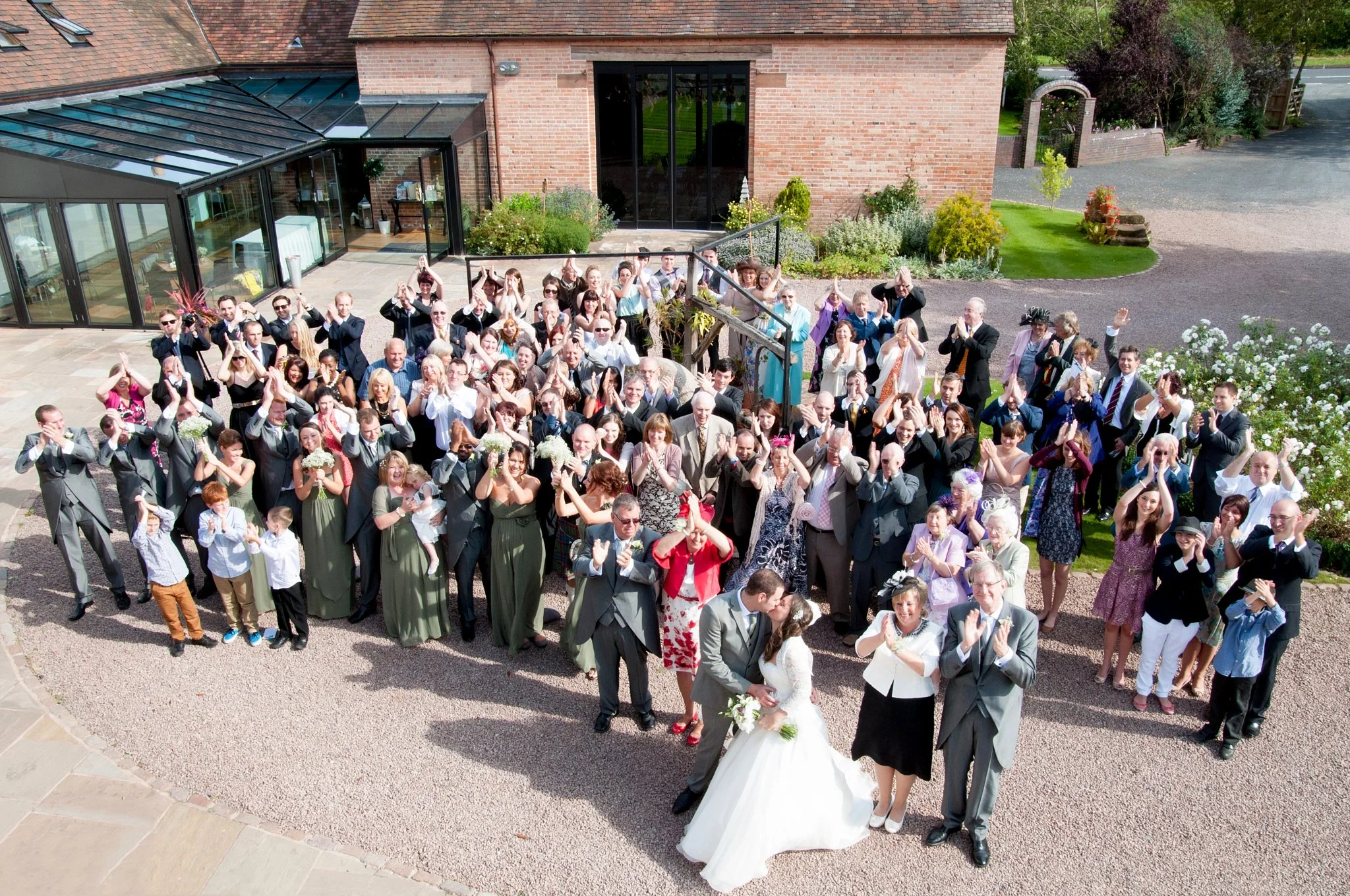 A large group of people gathered outside a brick building, celebrating a wedding, with some individuals clapping and waving, including a bride in a white dress and a groom in a suit.