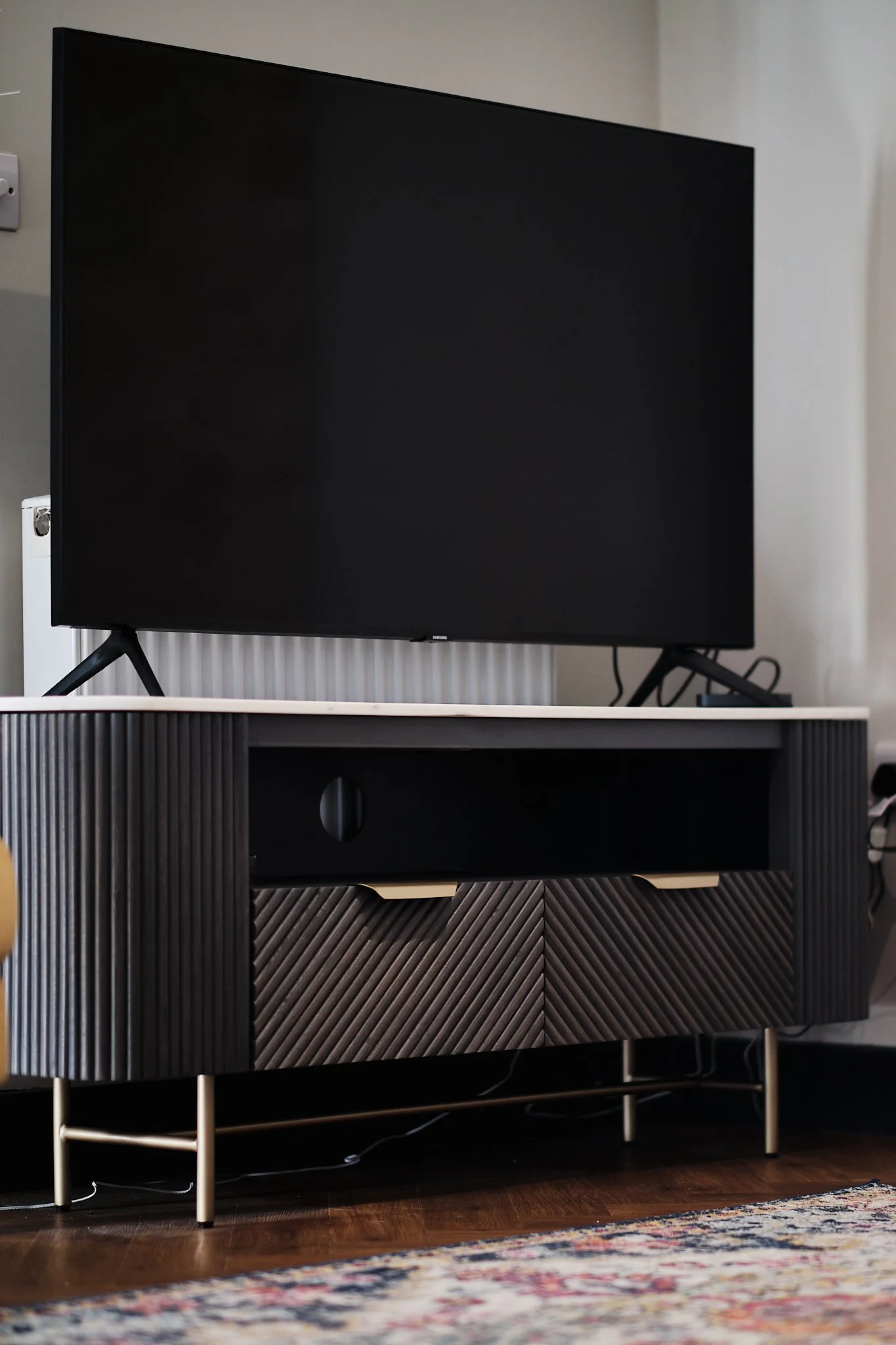 A flat-screen TV on a modern black and wood TV stand in a living room with hardwood floors and a patterned rug.