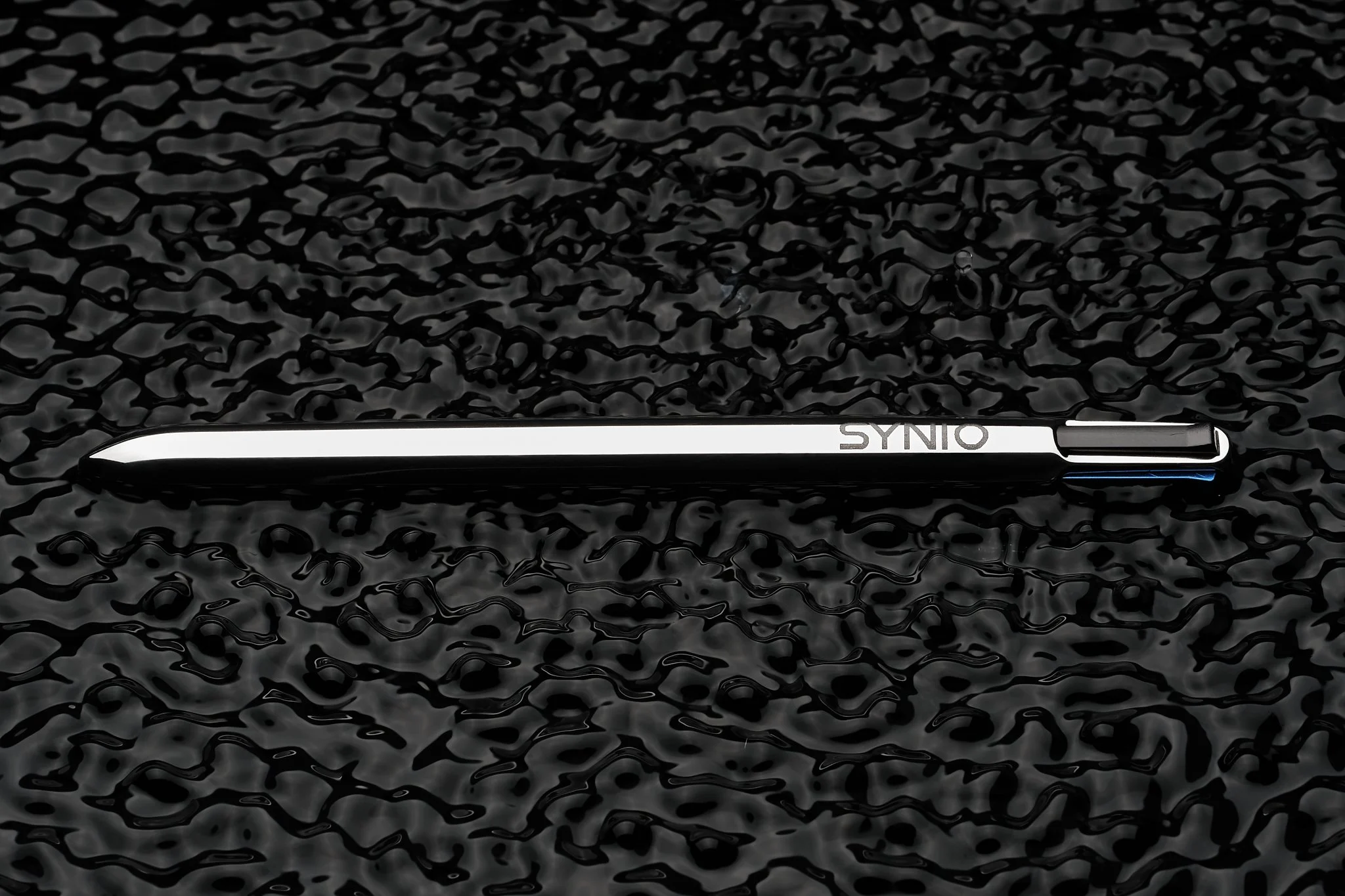 A black and silver stylus pen with the brand name 'SYNIO' on it, lying on a textured black surface.