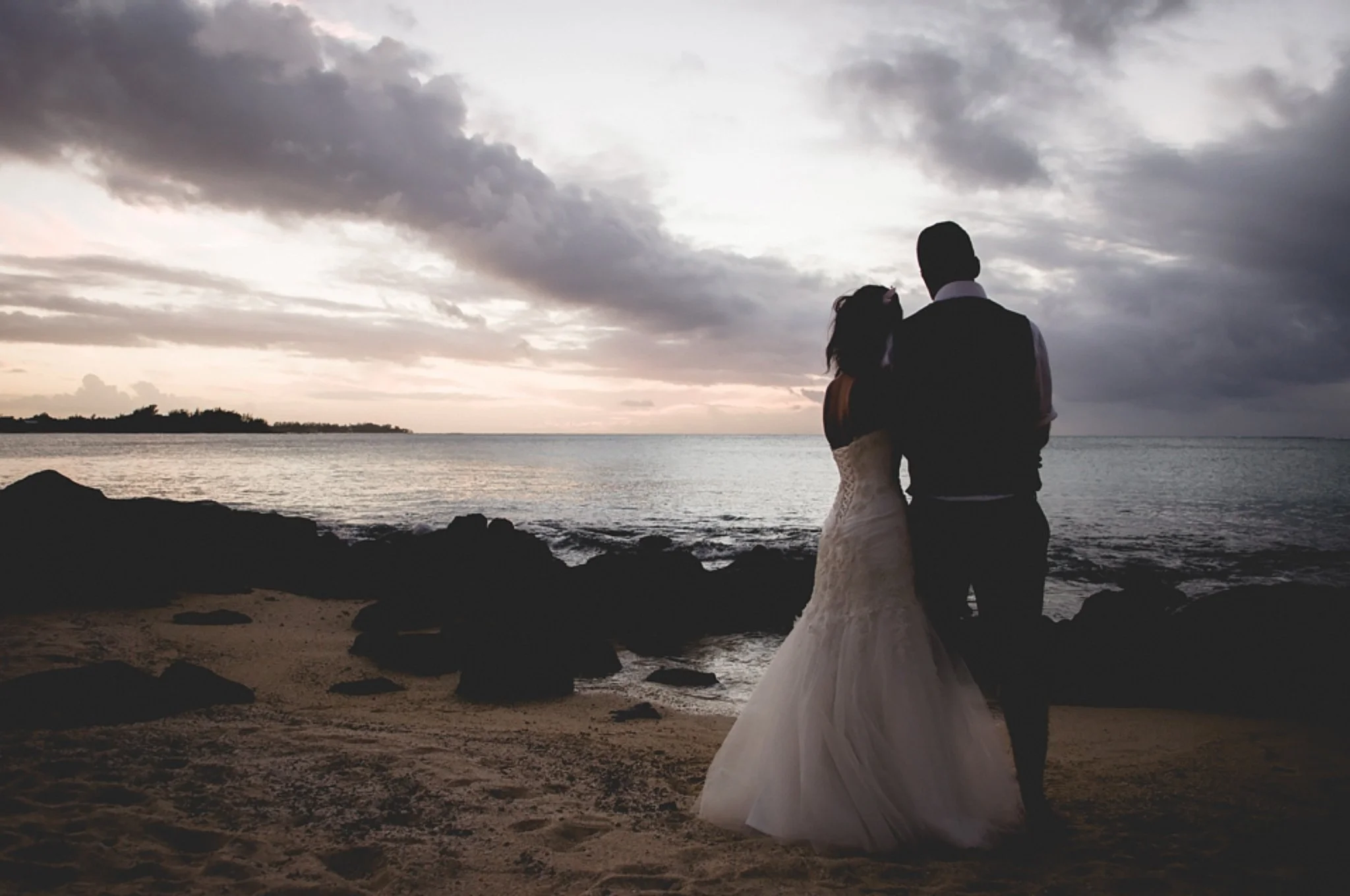A couple dressed in formal wedding attire standing on a beach at sunset, facing the ocean, with overcast skies overhead.