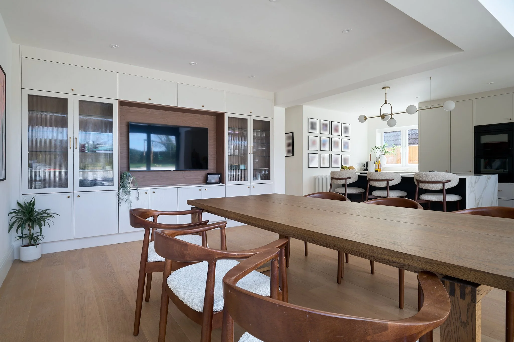 Modern open-concept living and dining area with white cabinetry, a wall-mounted TV, a large wooden dining table, wooden chairs, bar stools, and decorative framed art on the walls.