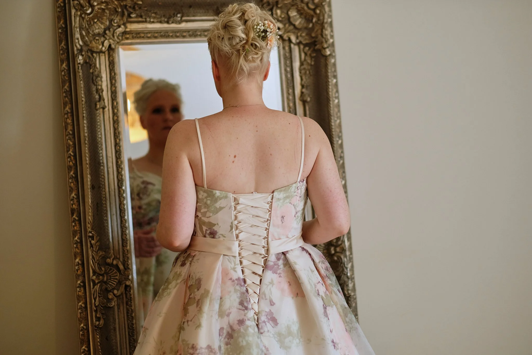 A woman with short, curly blonde hair adorned with small flowers is looking at her reflection in an ornate gold-framed mirror. She is wearing a pastel floral dress with a lace-up back. The setting appears to be a room with neutral walls.