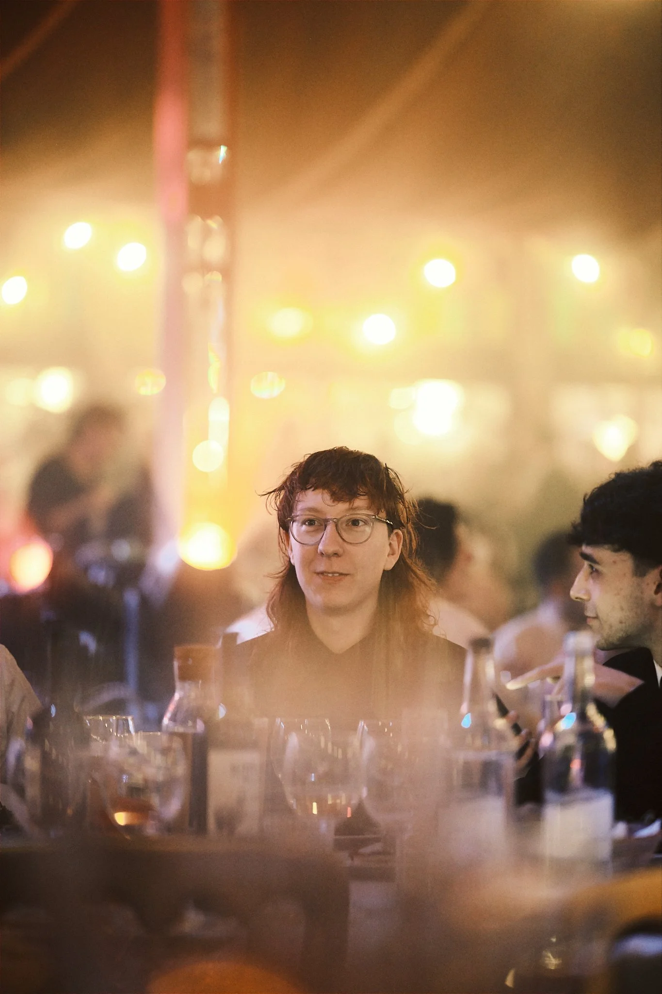 Person with glasses and long hair at a dinner party, surrounded by glasses and bottles, with warm, blurred string lights in the background.