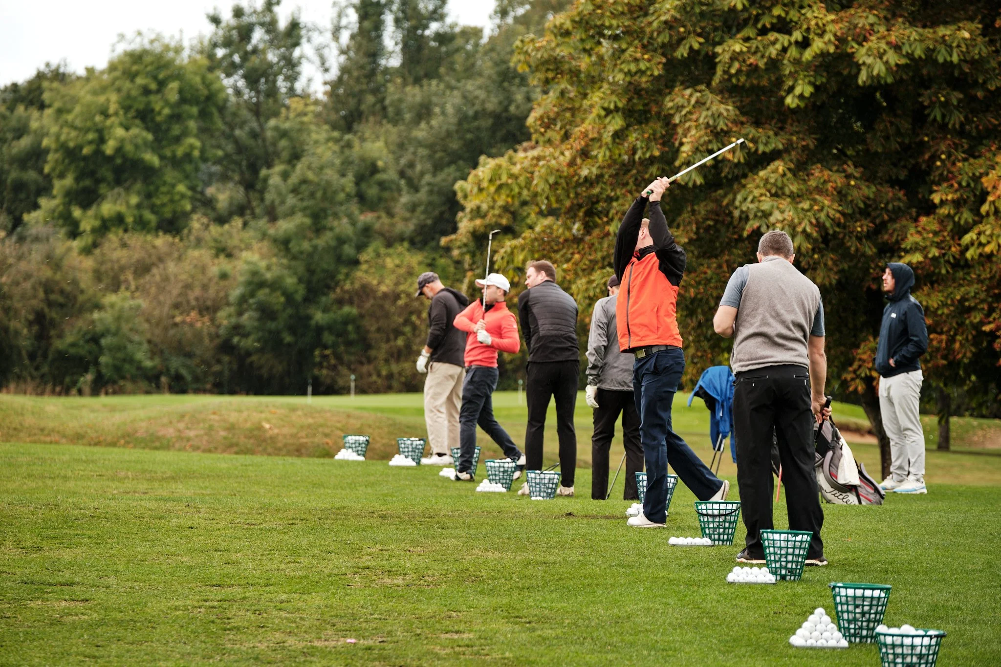 Several people practicing golf on a driving range, with baskets of golf balls and trees in the background.