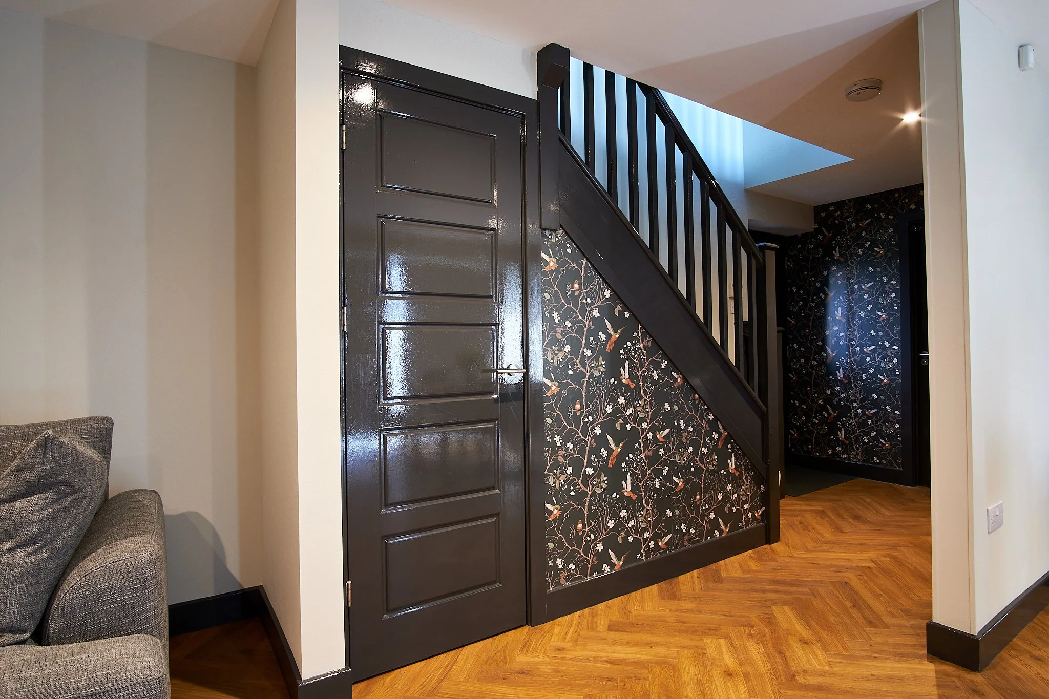 Interior view of a home showing a black staircase with a floral wallpaper underneath, a black door, and a beige wall on the left.