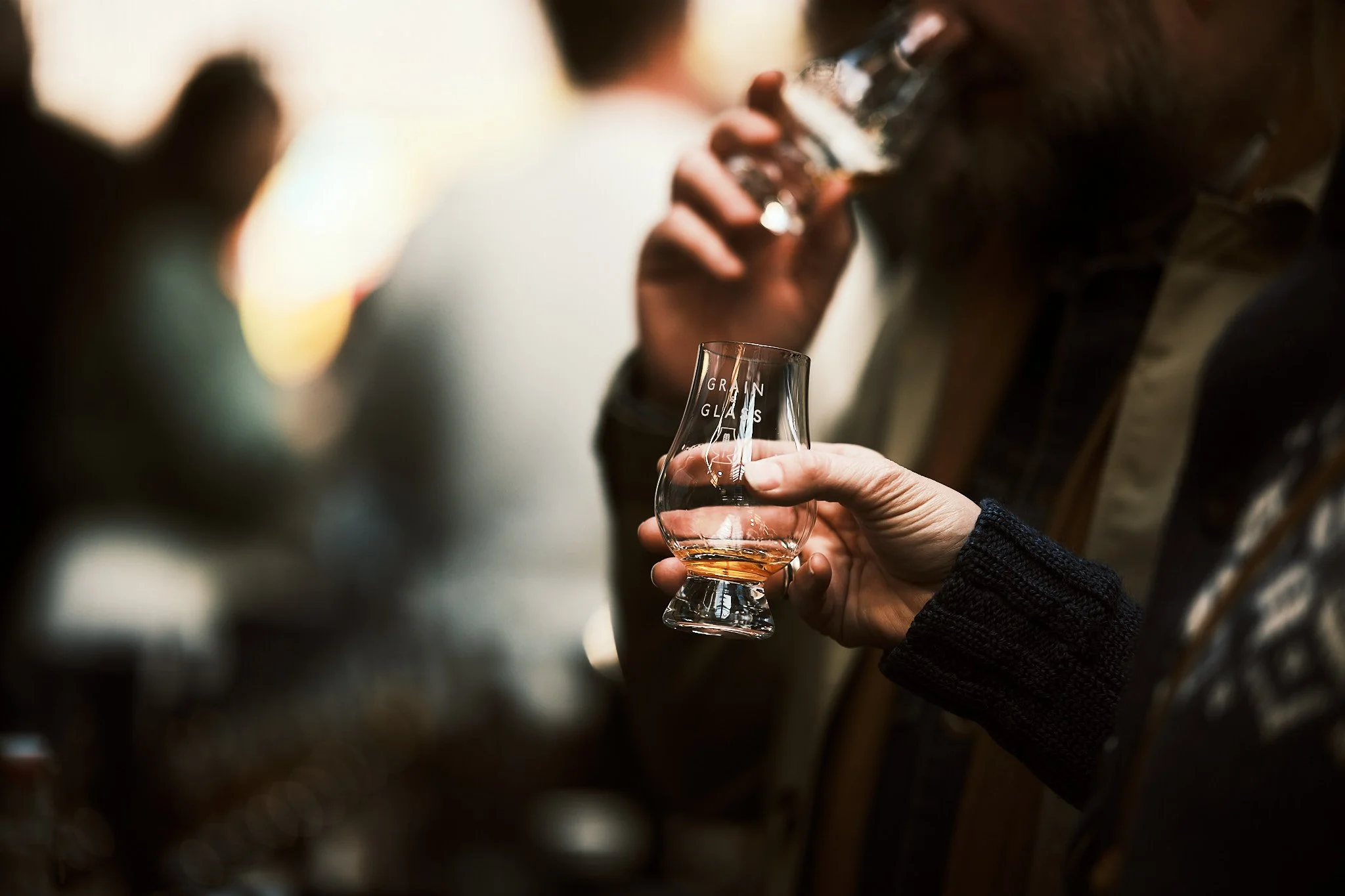 A person holding a glass with the words 'GRAIN GLASS' and drinking whiskey at a bar or social gathering, with another person in the background.
